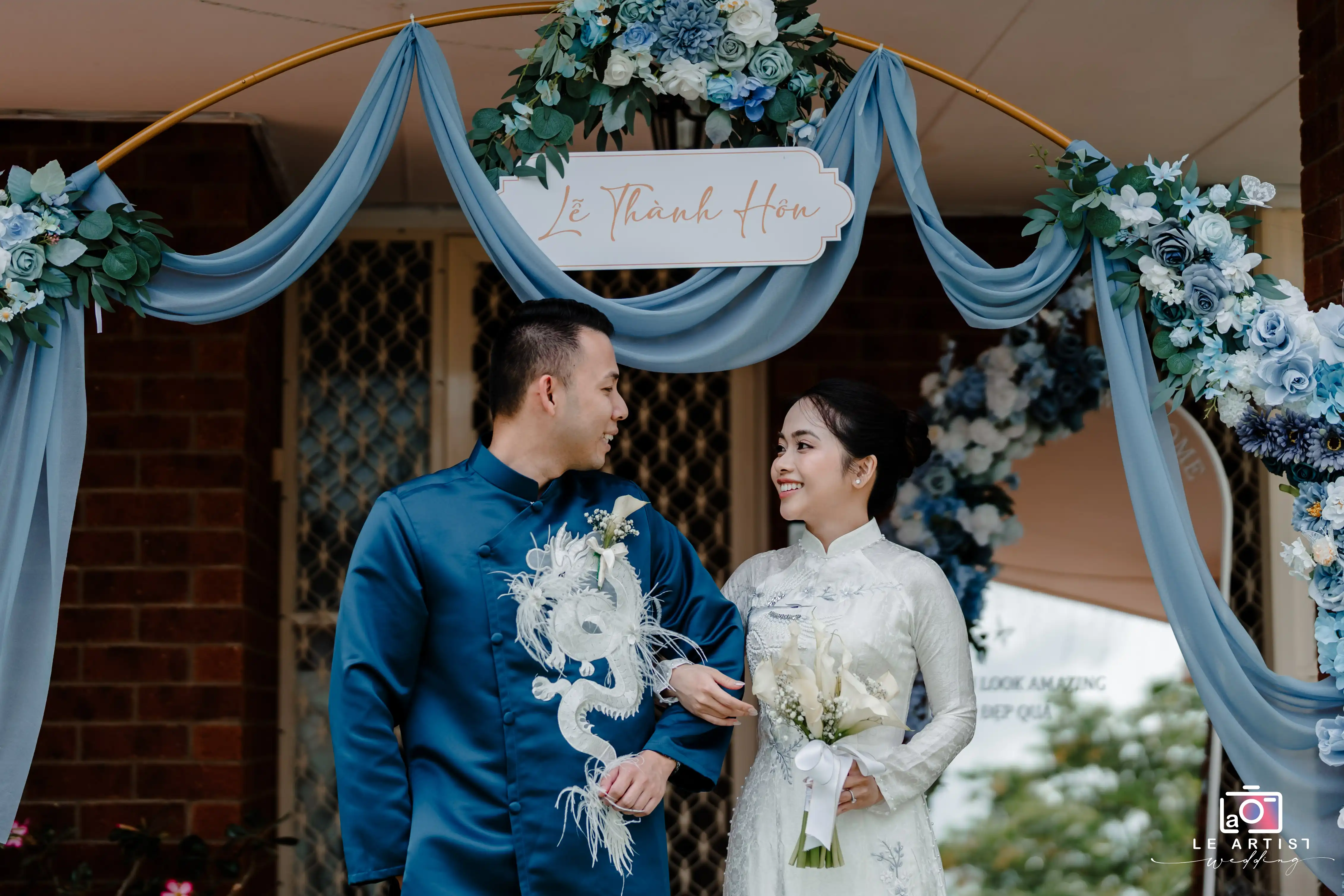 A joyful couple in traditional attire stands under a beautifully decorated archway, sharing a loving glance during their wedding celebration. Le Artist Wedding