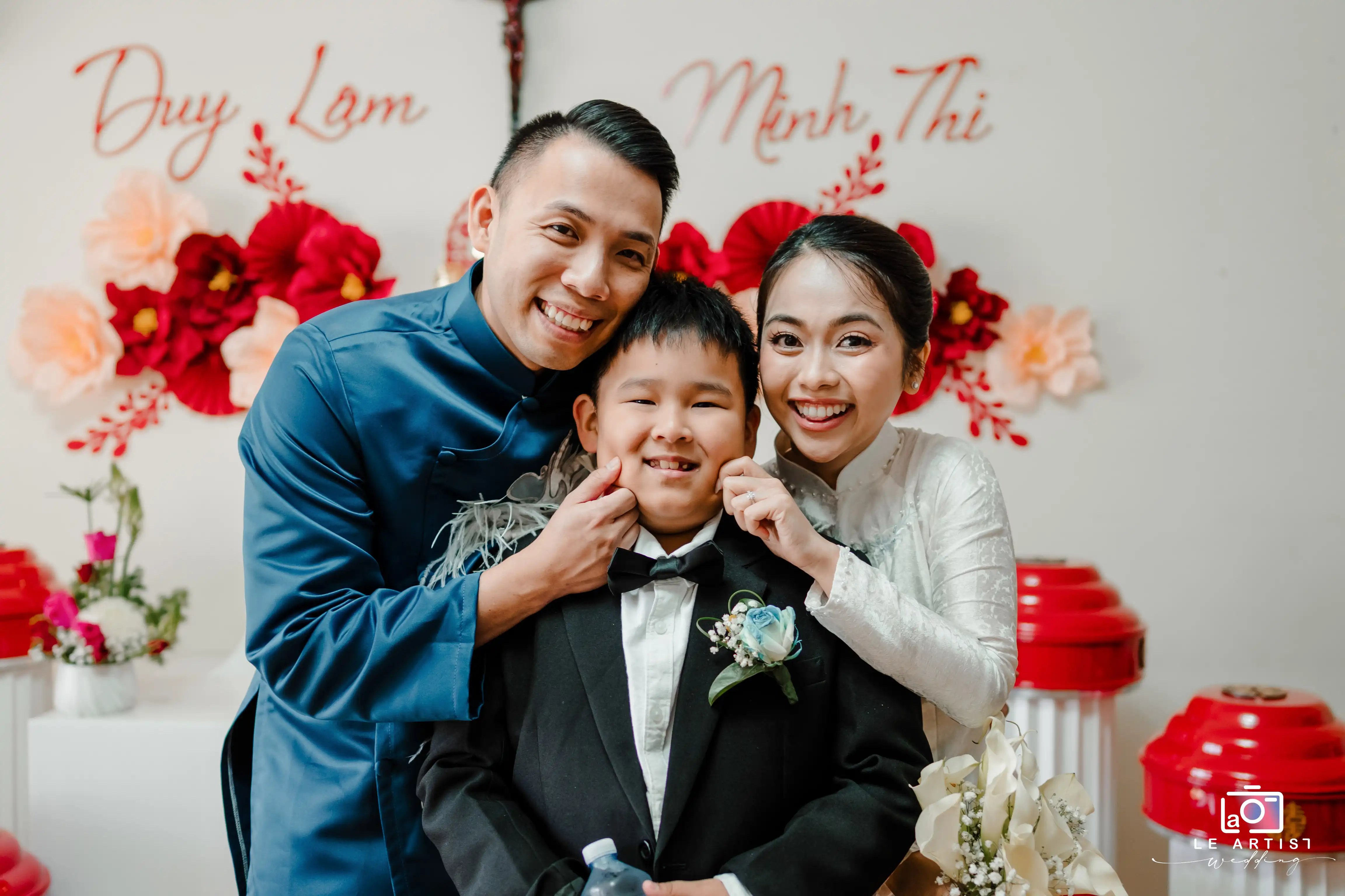 The couple joyfully poses with a young boy, creating a heartfelt family moment amidst vibrant floral decorations. Le Artist Wedding