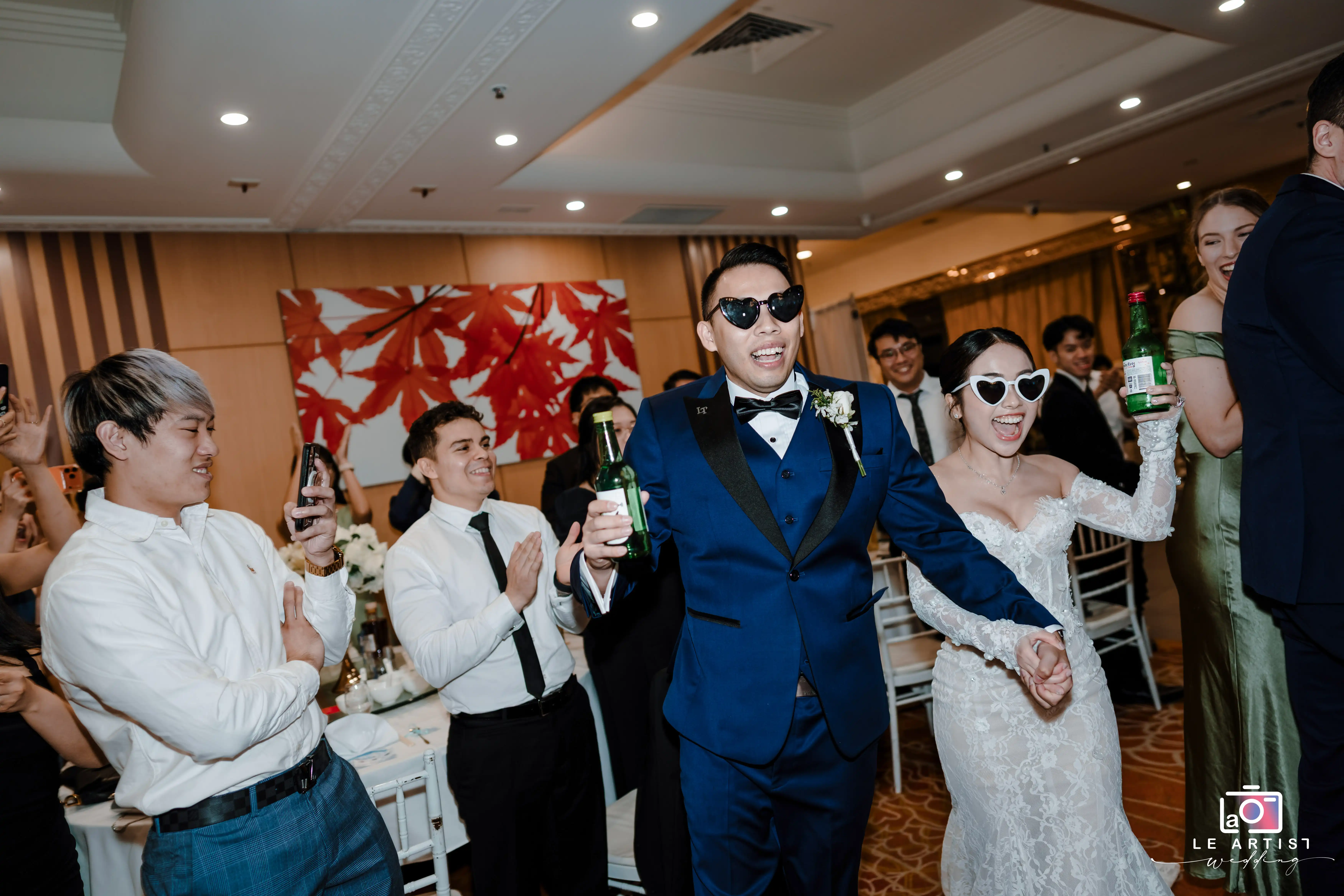 The newlyweds make a joyful entrance into their reception, wearing matching heart-shaped sunglasses and holding bottles, surrounded by cheering guests. Le Artist Wedding
