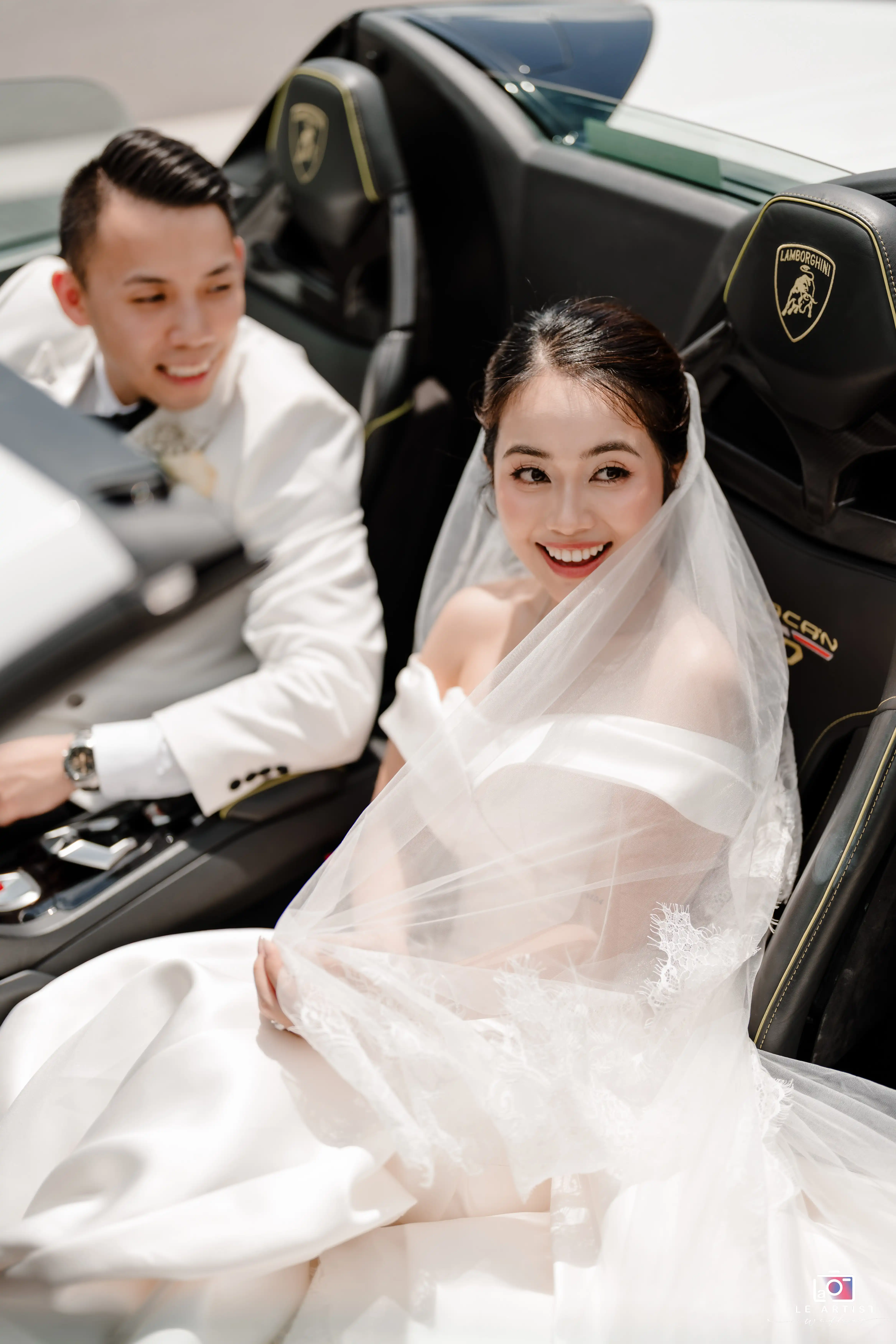 The newlyweds share a joyful moment seated in a luxurious car, with the bride's veil delicately draped around her, capturing a sense of elegance and excitement. Le Artist Wedding