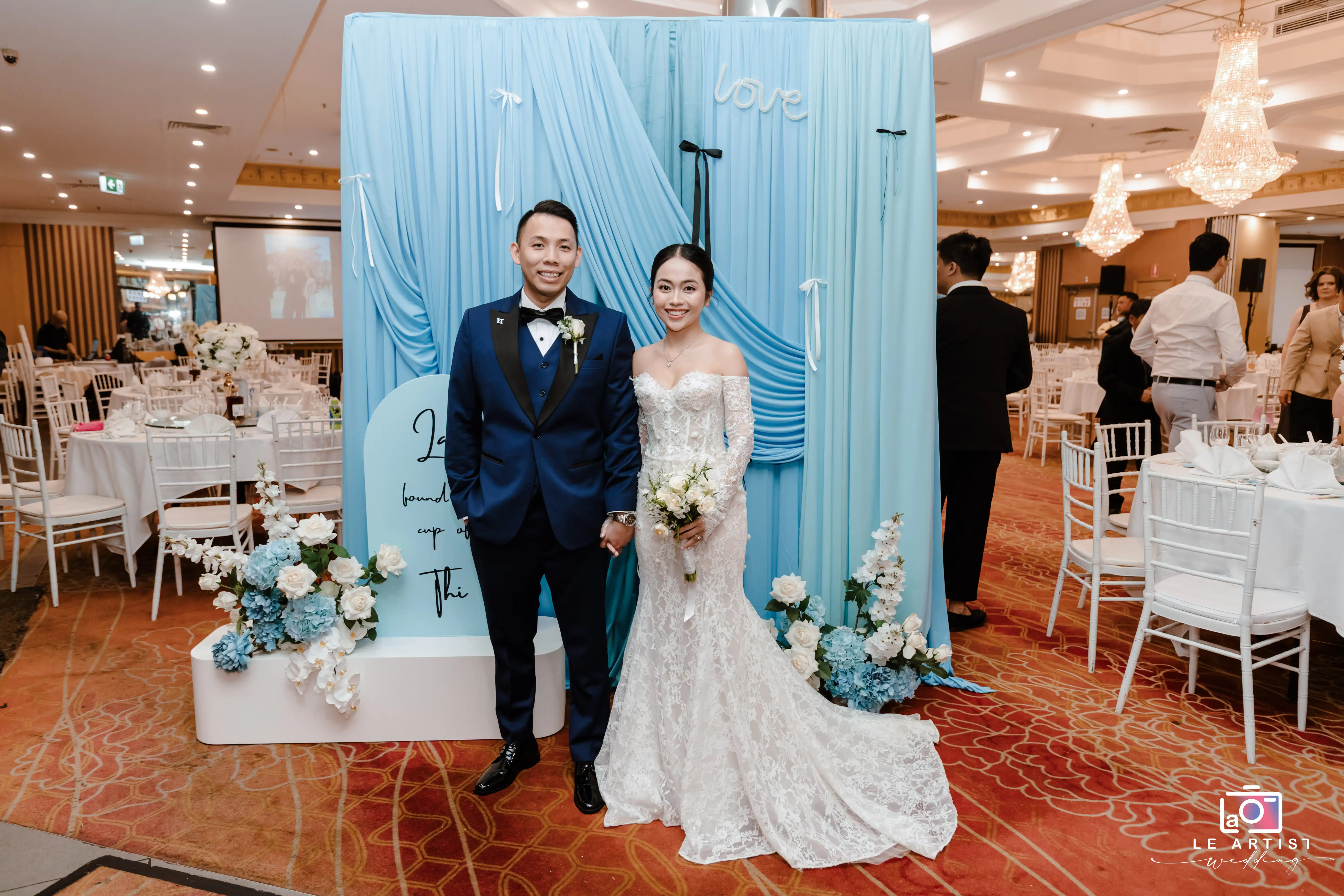 The newlyweds pose with satisfaction in front of a beautifully decorated backdrop, embodying the joy of their wedding celebration. Le Artist Wedding