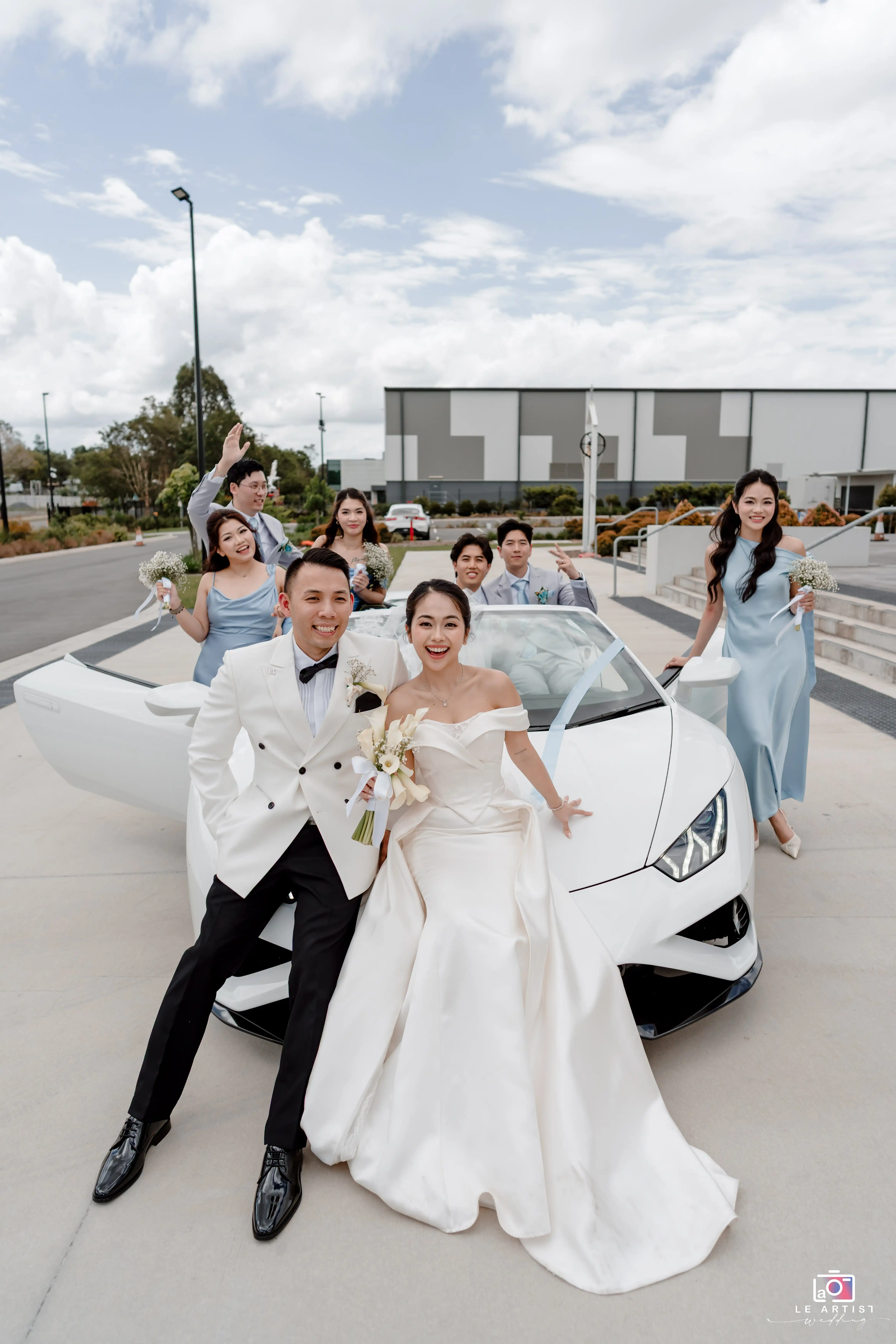 The newlyweds joyfully pose with their wedding party in front of a sleek white car, capturing a vibrant and celebratory moment. Le Artist Wedding