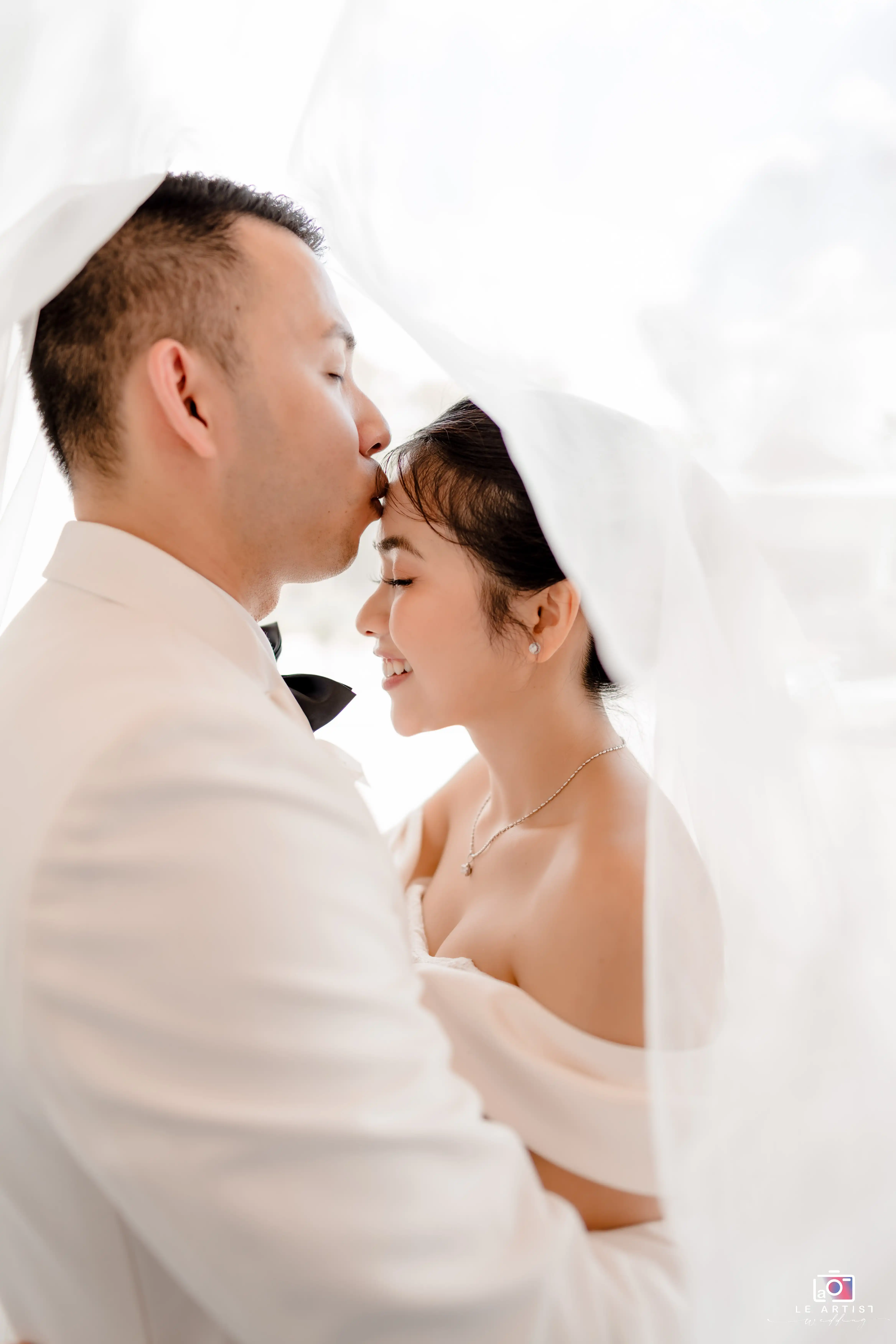 A tender moment under the veil as the groom gently kisses the bride's forehead, capturing their intimate connection. Le Artist Wedding
