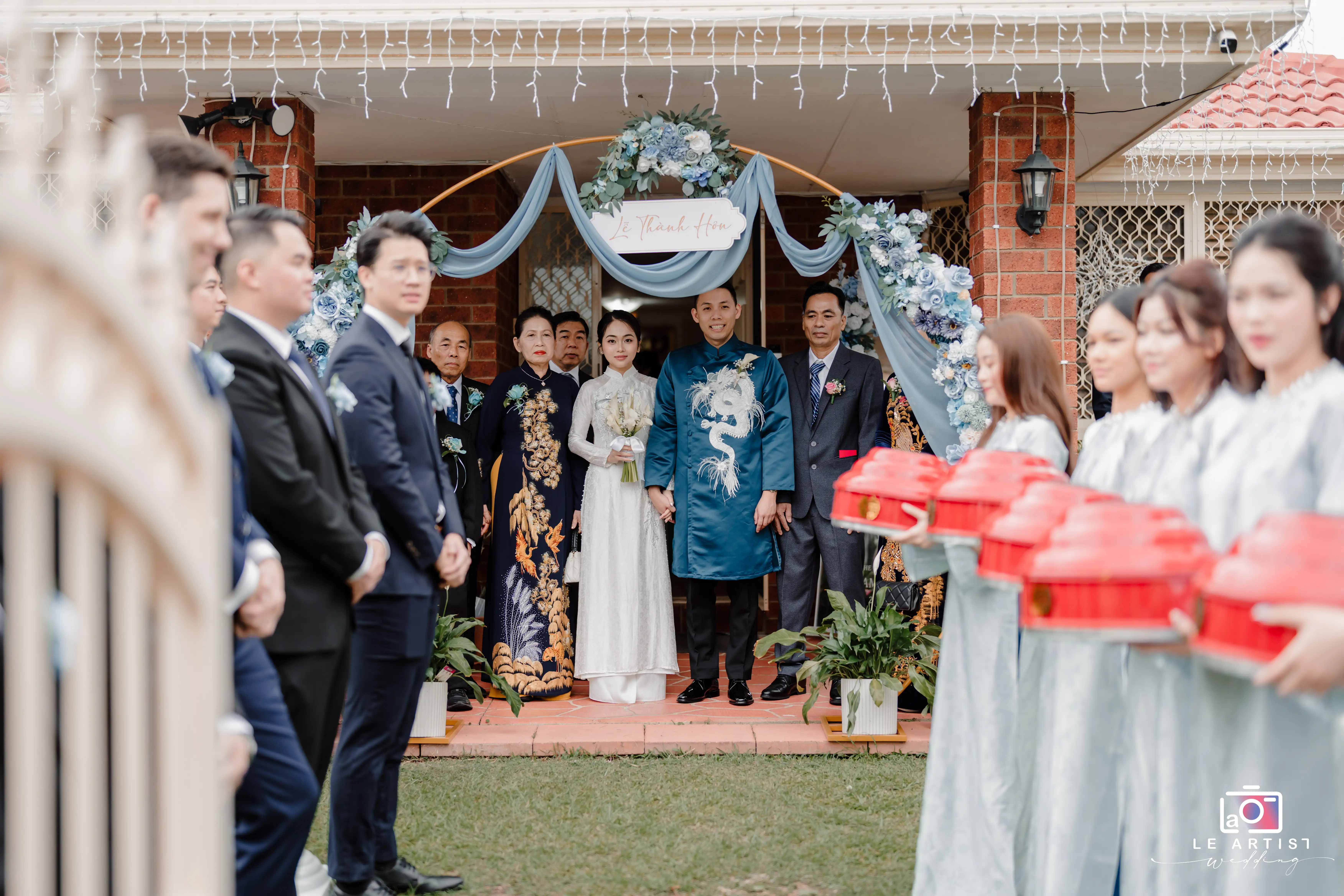 The couple poses with family and guests in traditional attire during a Vietnamese wedding ceremony, surrounded by an elegantly decorated floral archway. Le Artist Wedding