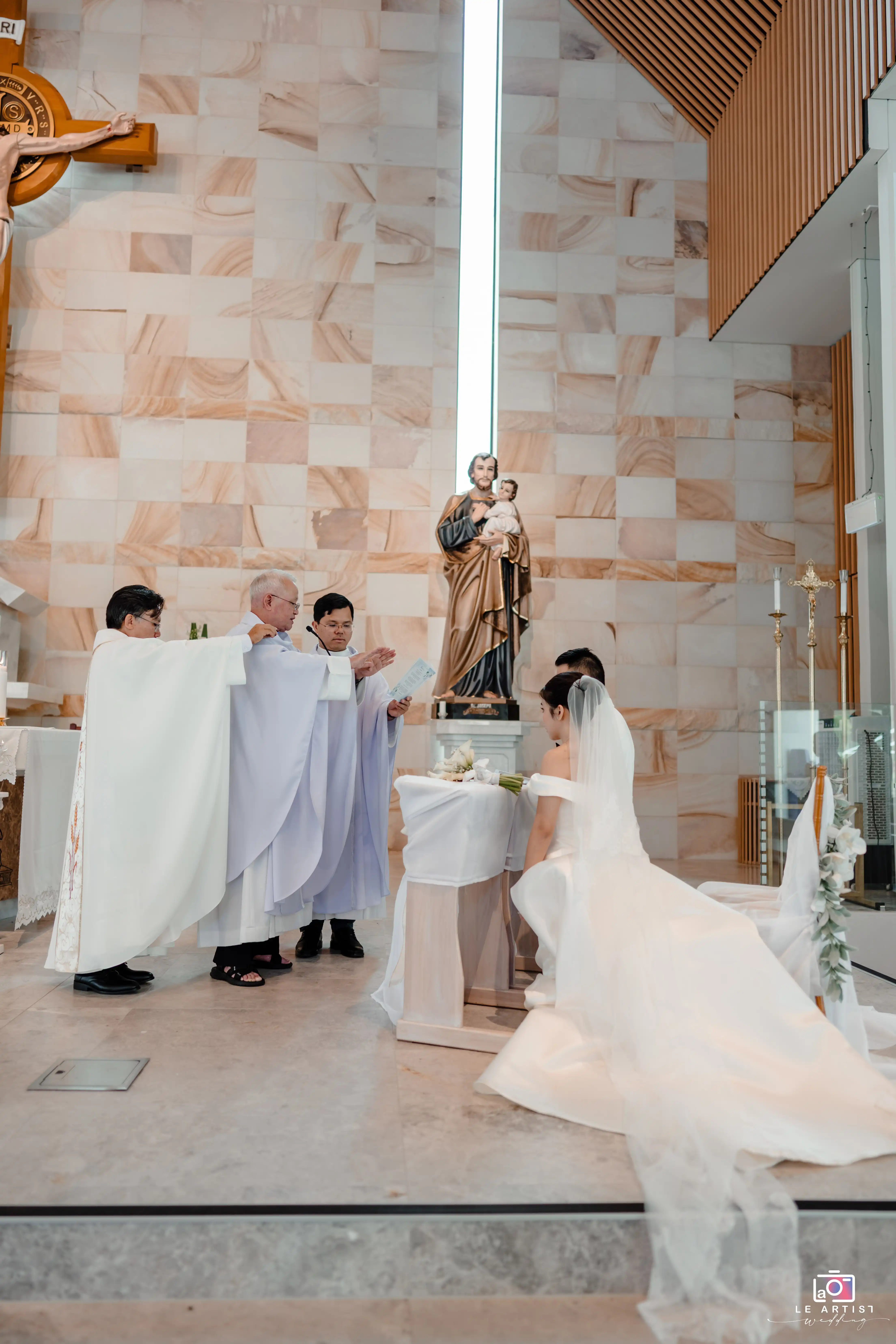 The couple kneels at the altar during a solemn church ceremony, surrounded by clergy performing a blessing. The setting features a beautifully adorned church interior. Le Artist Wedding