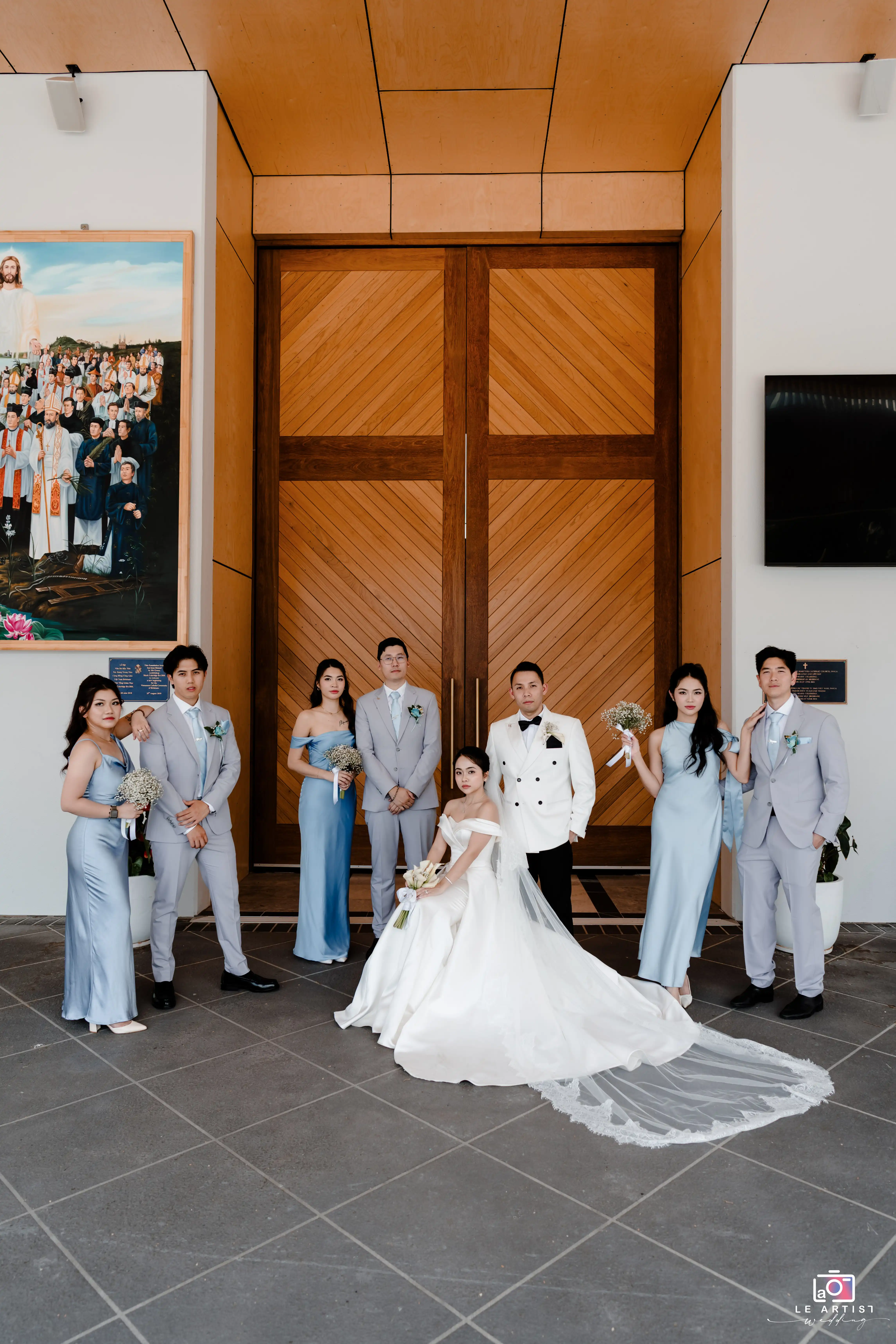 The bride and groom pose elegantly with their bridal party in front of grand wooden doors, showcasing their finely coordinated attire in soothing shades against the church's ornate setting. Le Artist Wedding
