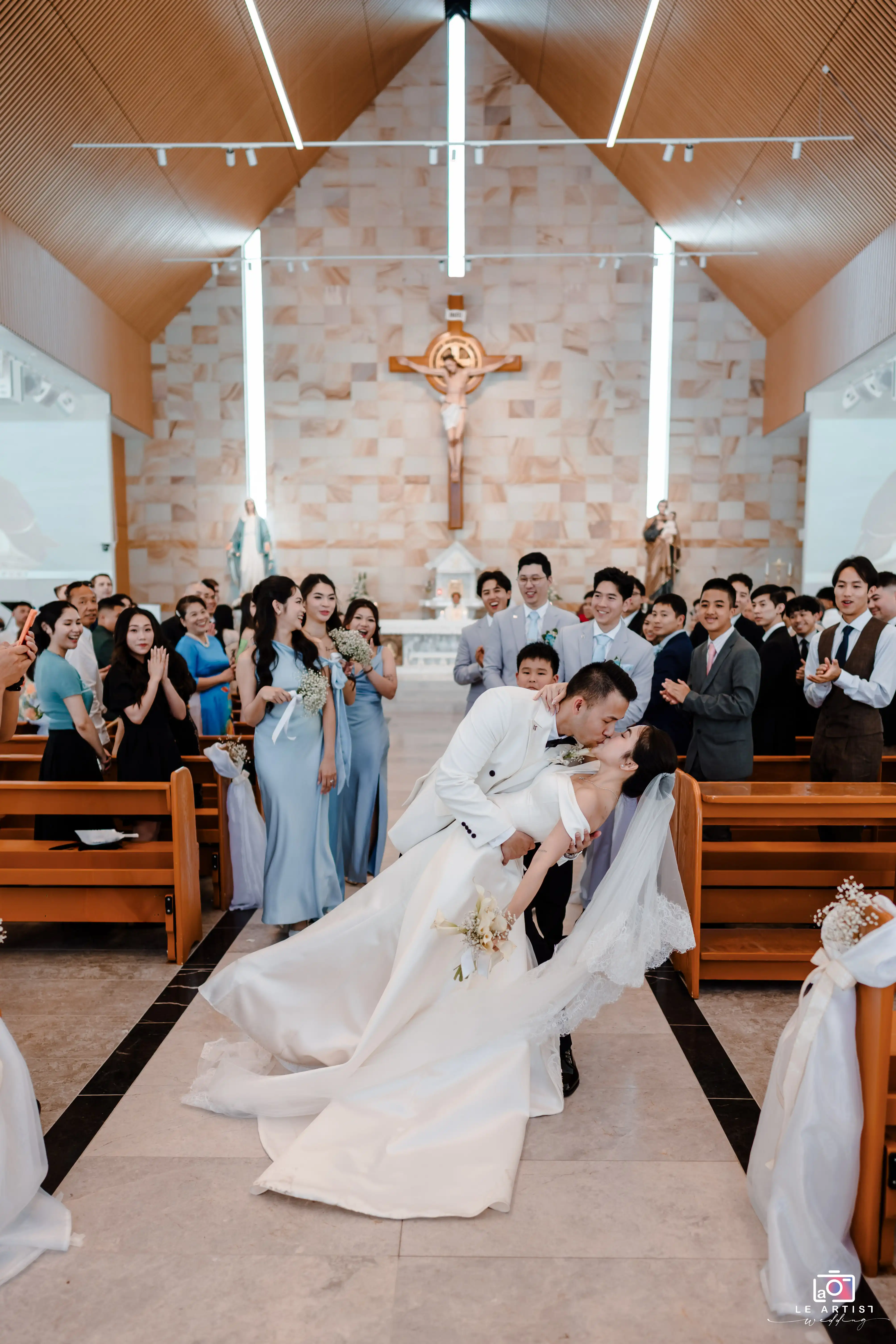 The newlyweds share a passionate kiss in the church, surrounded by joyful friends and family applauding their union. Le Artist Wedding