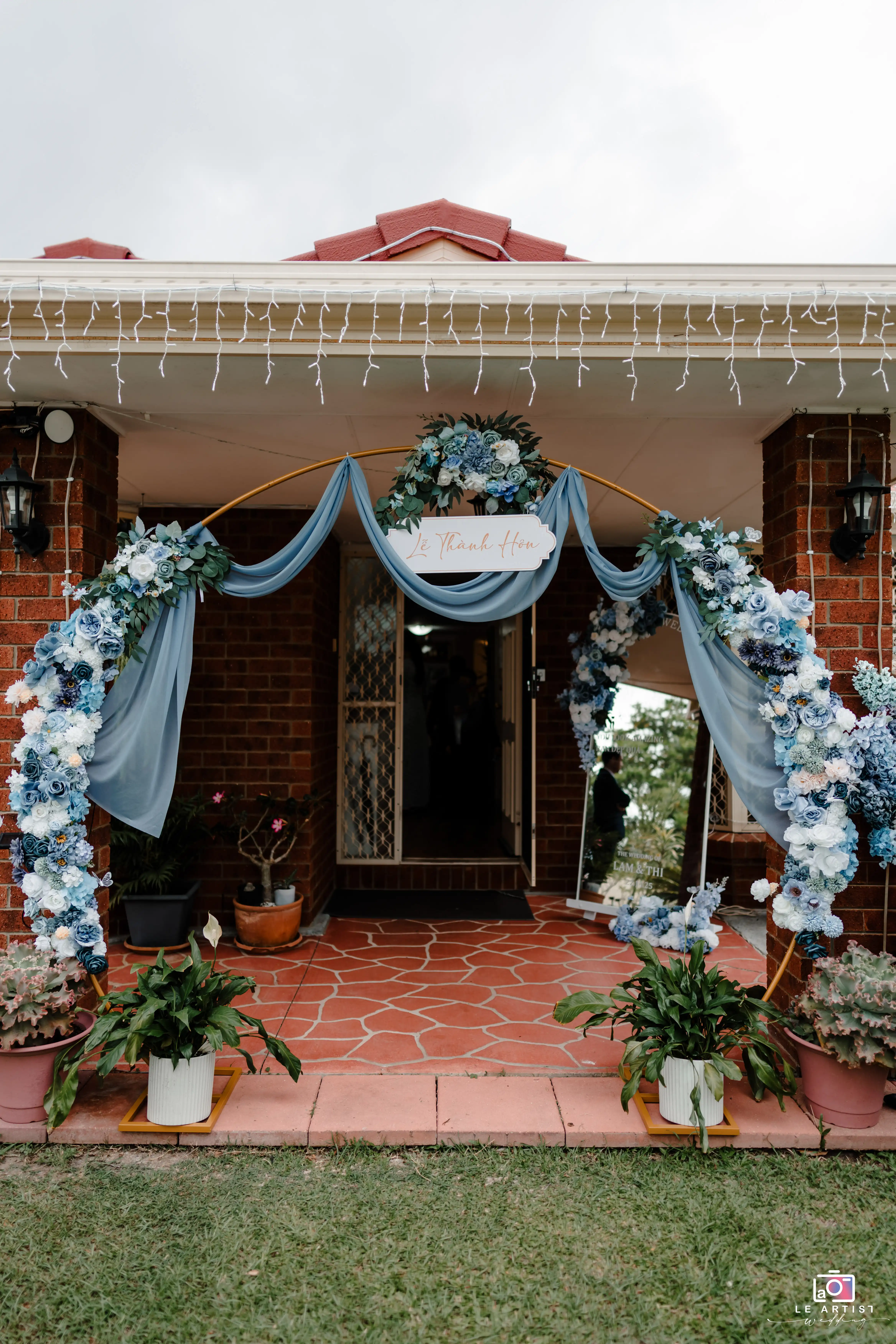 An elegantly decorated entrance with a flower arch and draped fabric in soothing blue tones, setting a welcoming atmosphere for the wedding ceremony. Le Artist Wedding