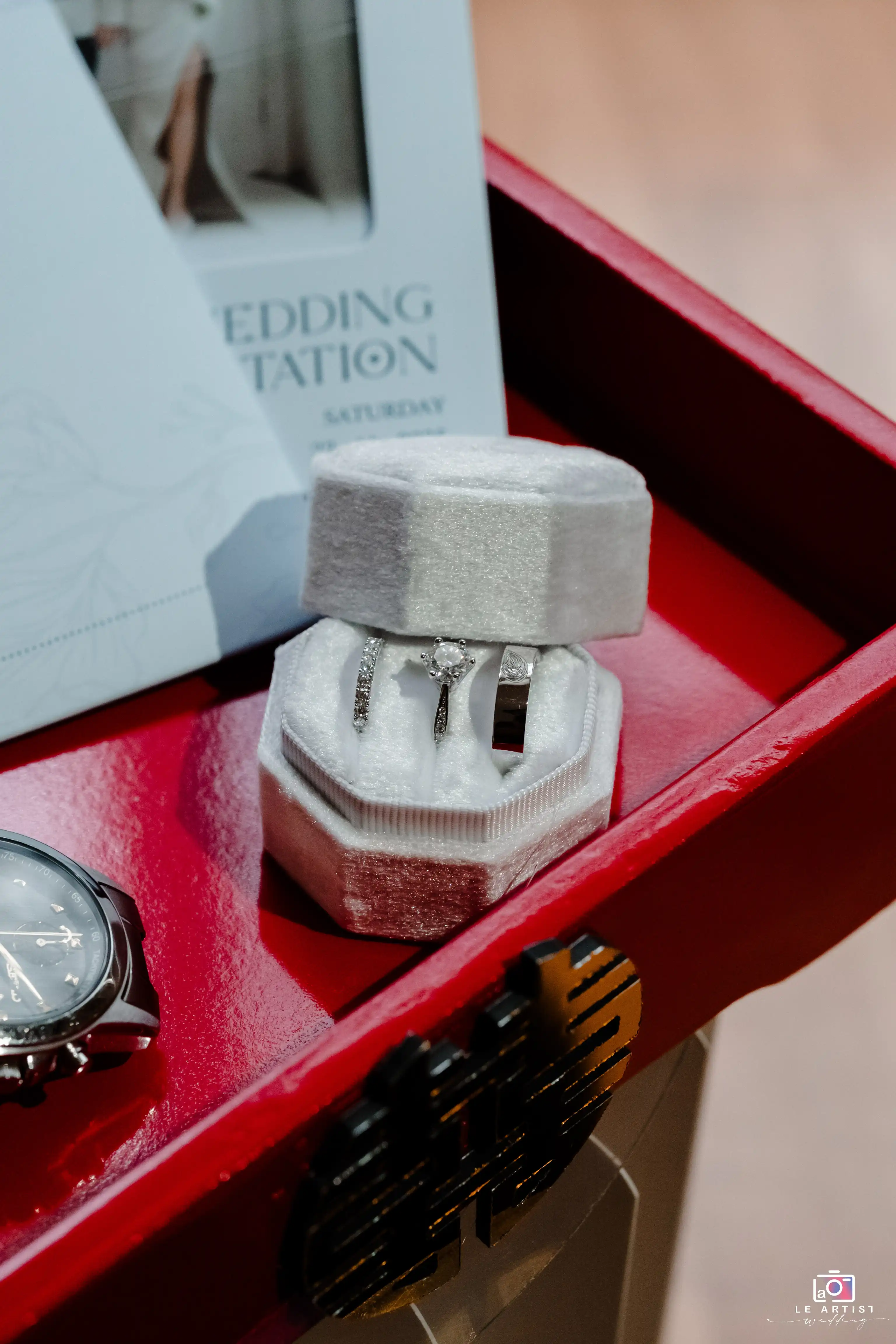 A beautifully arranged gallery display featuring the wedding rings in an elegant velvet box alongside a classic watch and wedding invitation, set in a vibrant red tray with traditional motifs. Le Artist Wedding