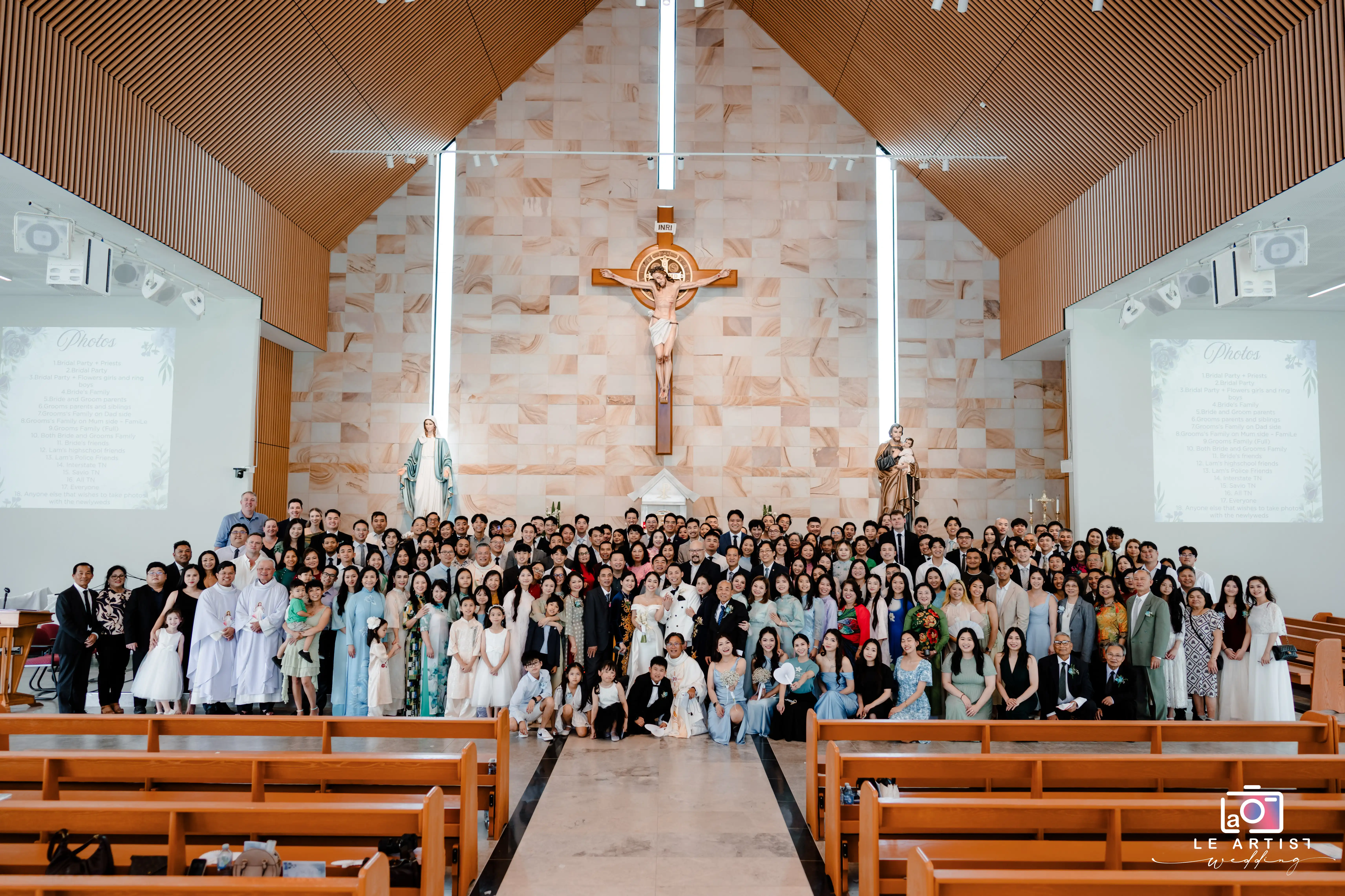 The newlyweds pose with their family and friends for a group photo in a beautifully adorned church, capturing a memorable moment of unity and celebration. Le Artist Wedding