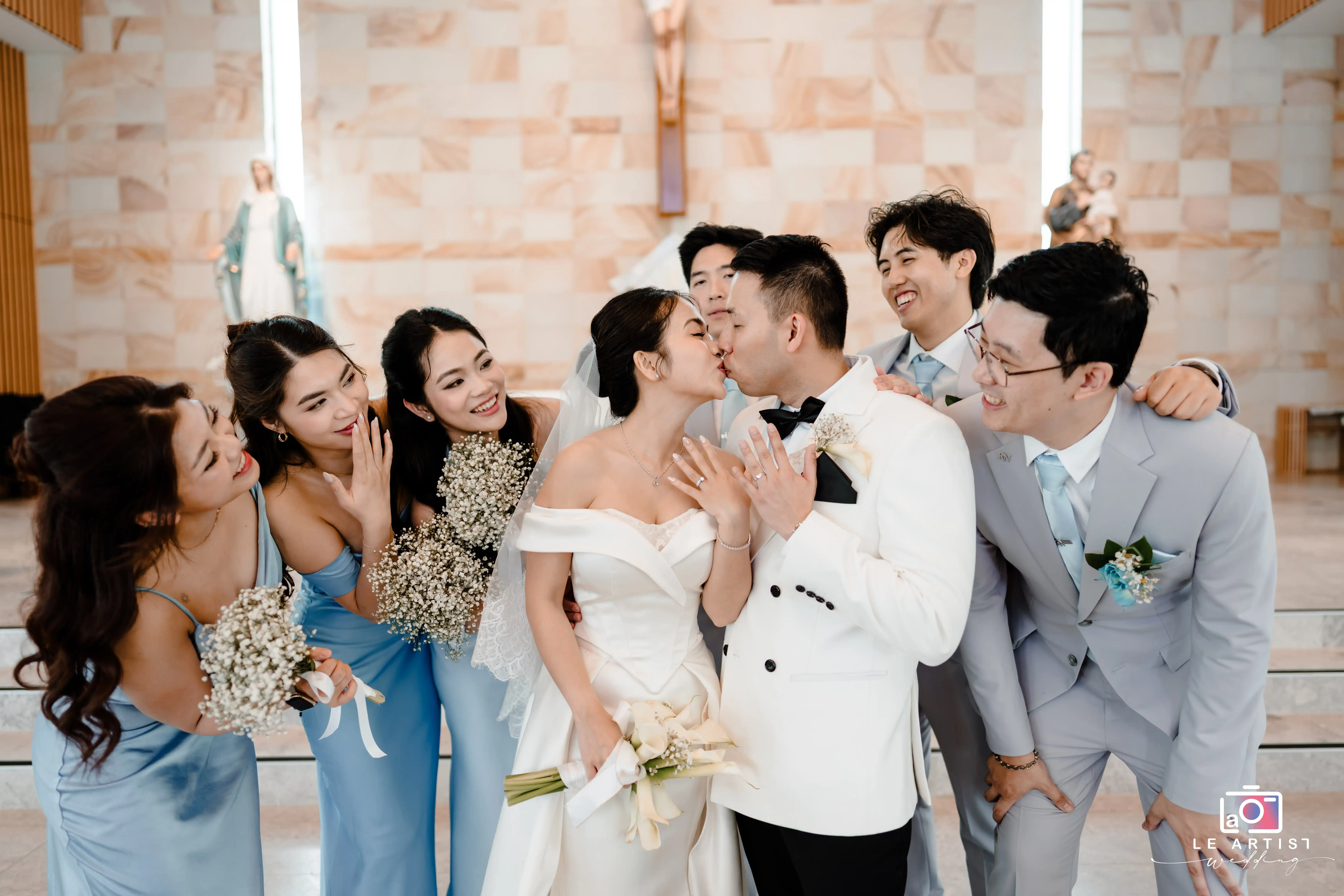 The joyful wedding party celebrates the newlyweds' kiss, with bridesmaids in matching blue dresses and groomsmen in light suits sharing in the newlyweds' happiness. Le Artist Wedding