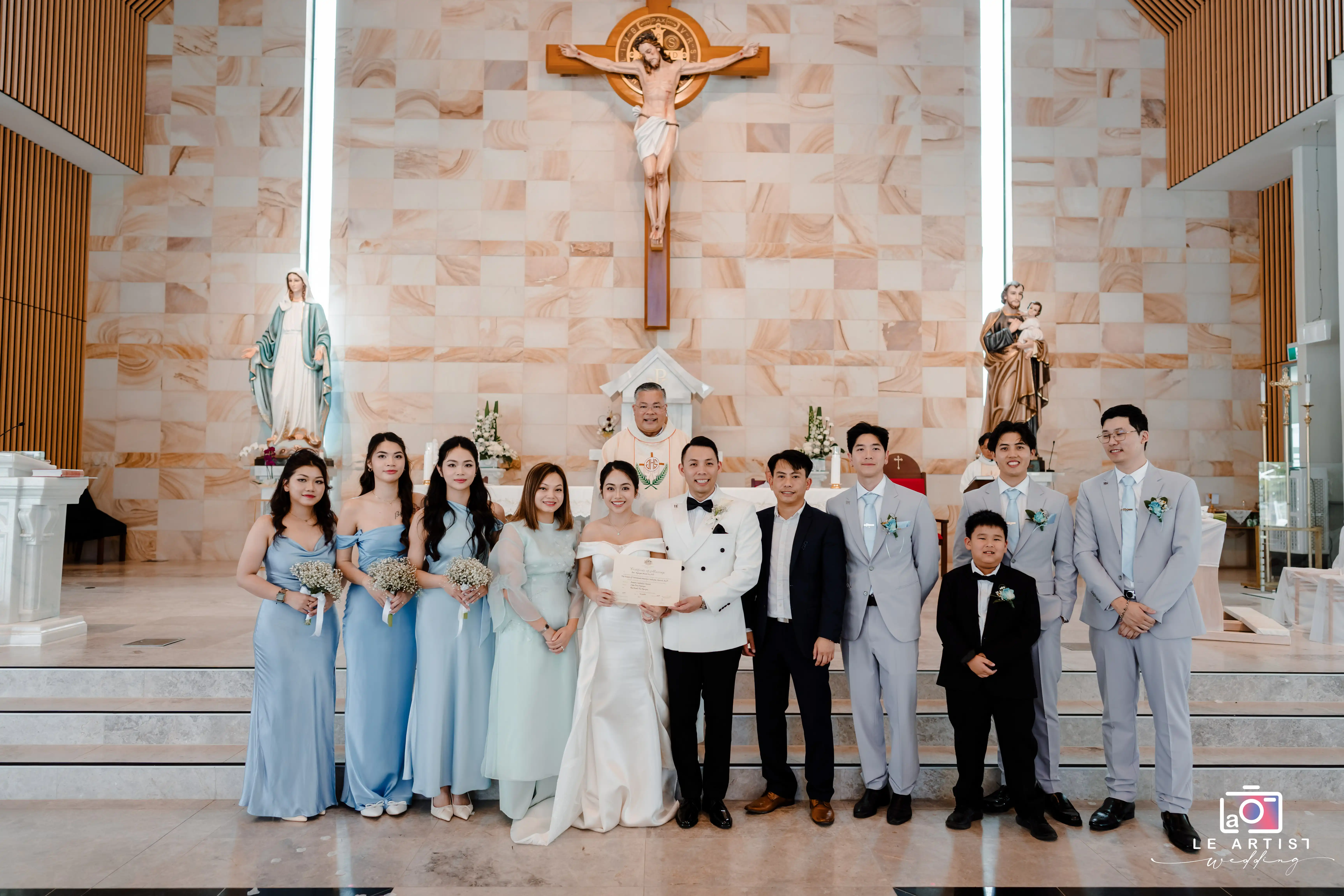 The wedding party gathers in the church for a group photo, showcasing a beautifully coordinated blend of blue dresses and light gray suits. The newlyweds hold their marriage certificate, standing proudly at the altar surrounded by friends and family. Le Artist Wedding
