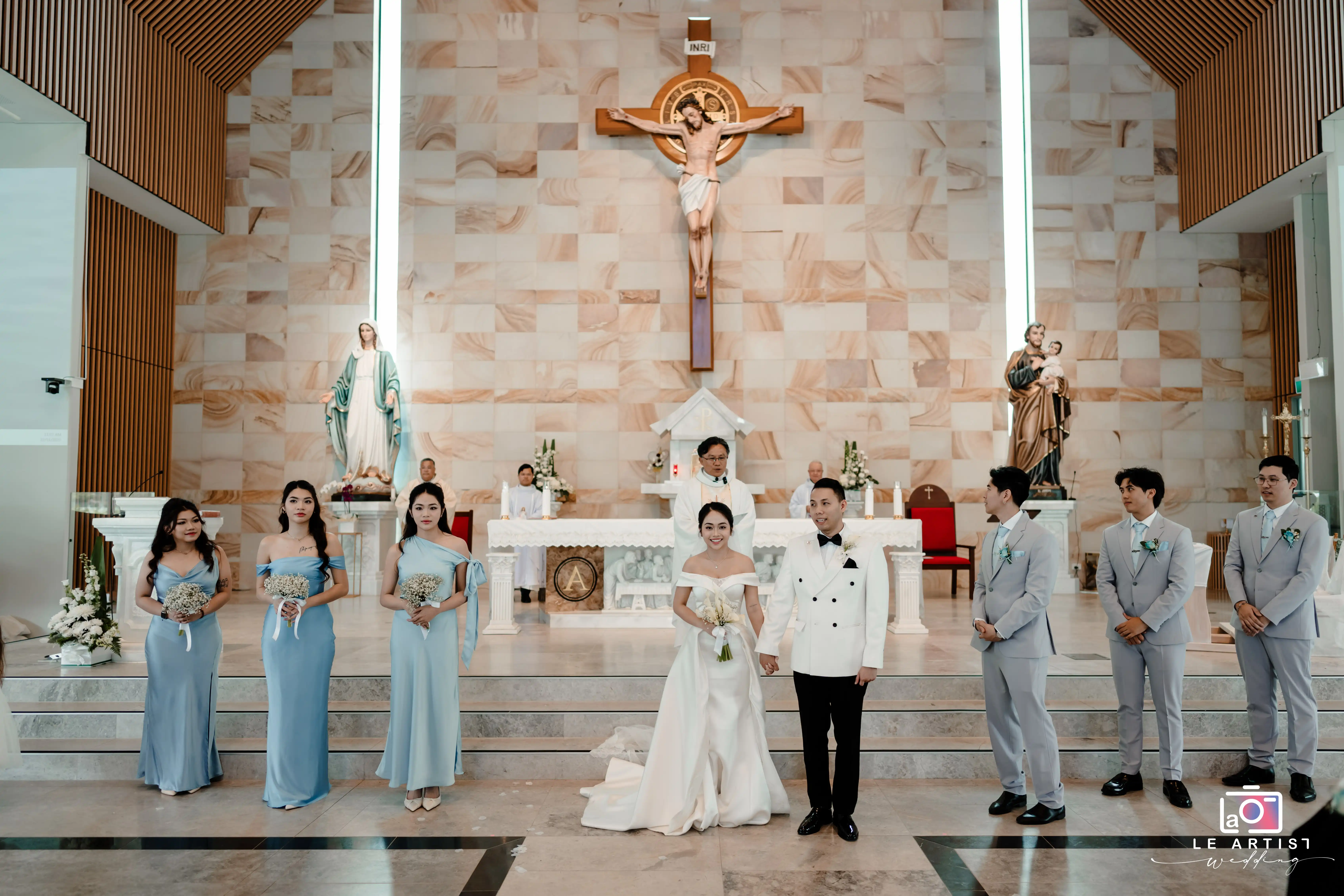 The wedding party stands together in a beautifully adorned church, with the bridesmaids and groomsmen dressed in elegant attire, complementing the bride and groom who are joyfully holding hands at the altar. Le Artist Wedding