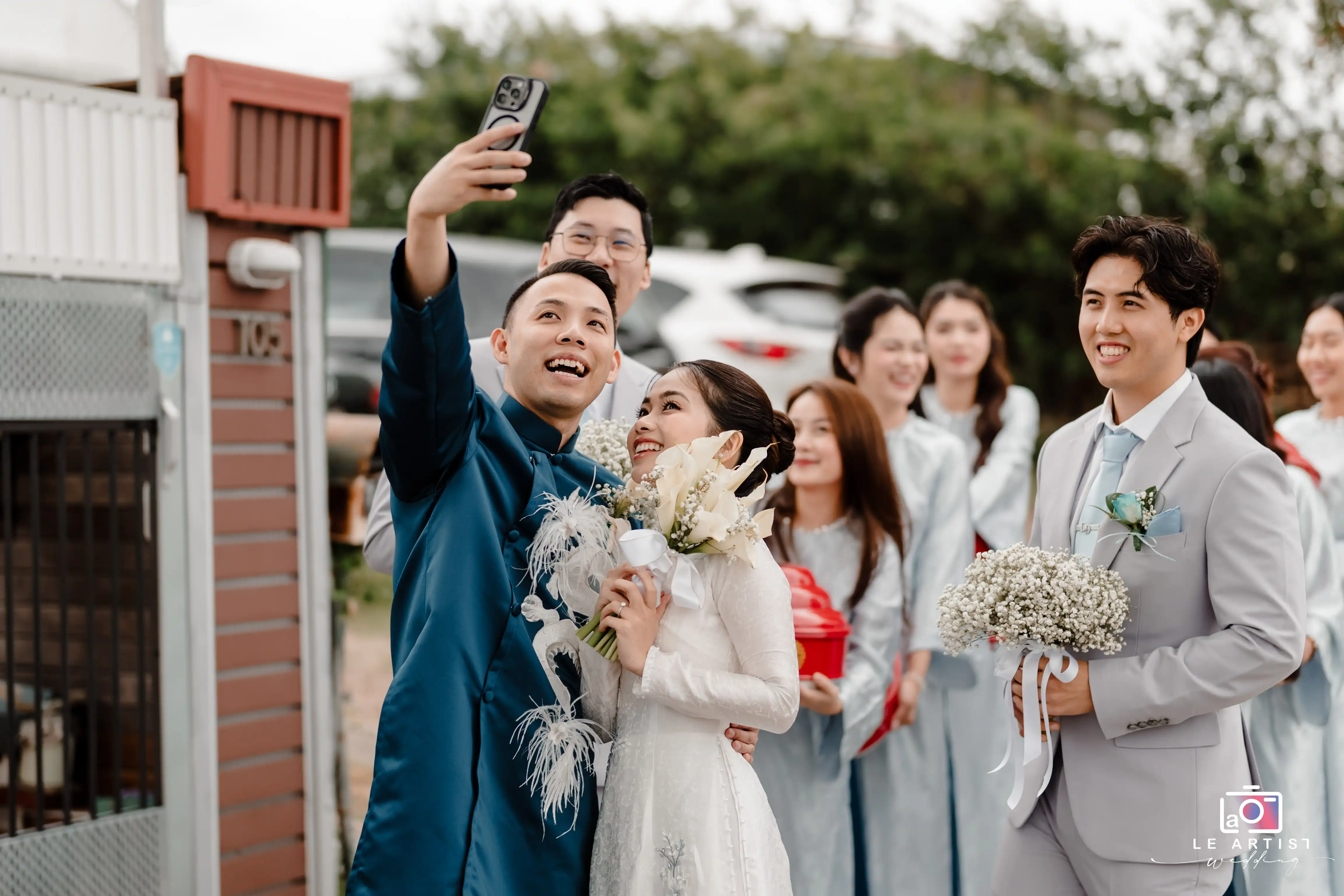 The wedding party joyfully gathers for a selfie, capturing the moment with smiles and flowers, while celebrating the special day together. Le Artist Wedding