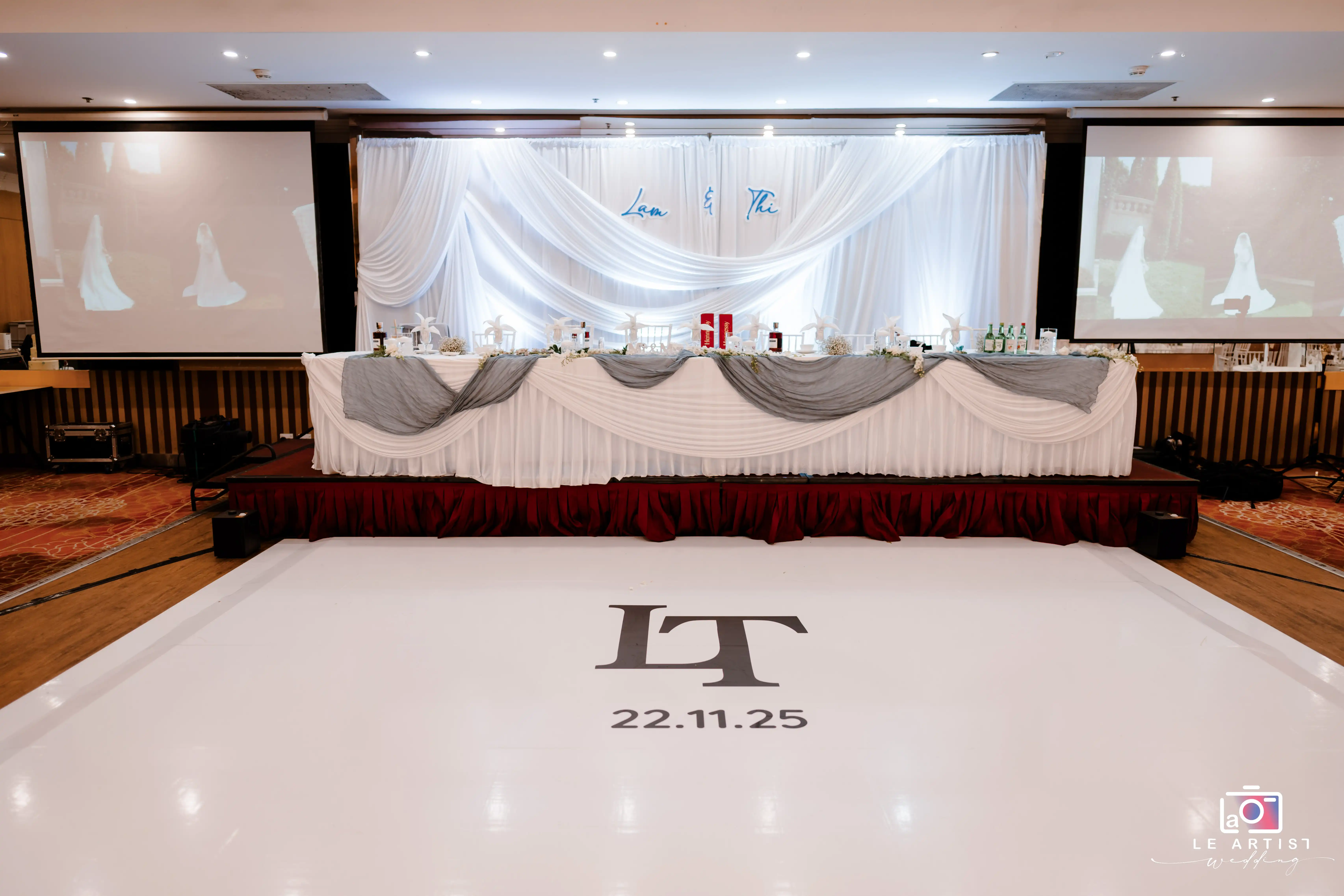 Elegant wedding reception setup featuring a beautifully decorated sweetheart table with a white and grey color scheme, flanked by projectors displaying wedding images. Le Artist Wedding