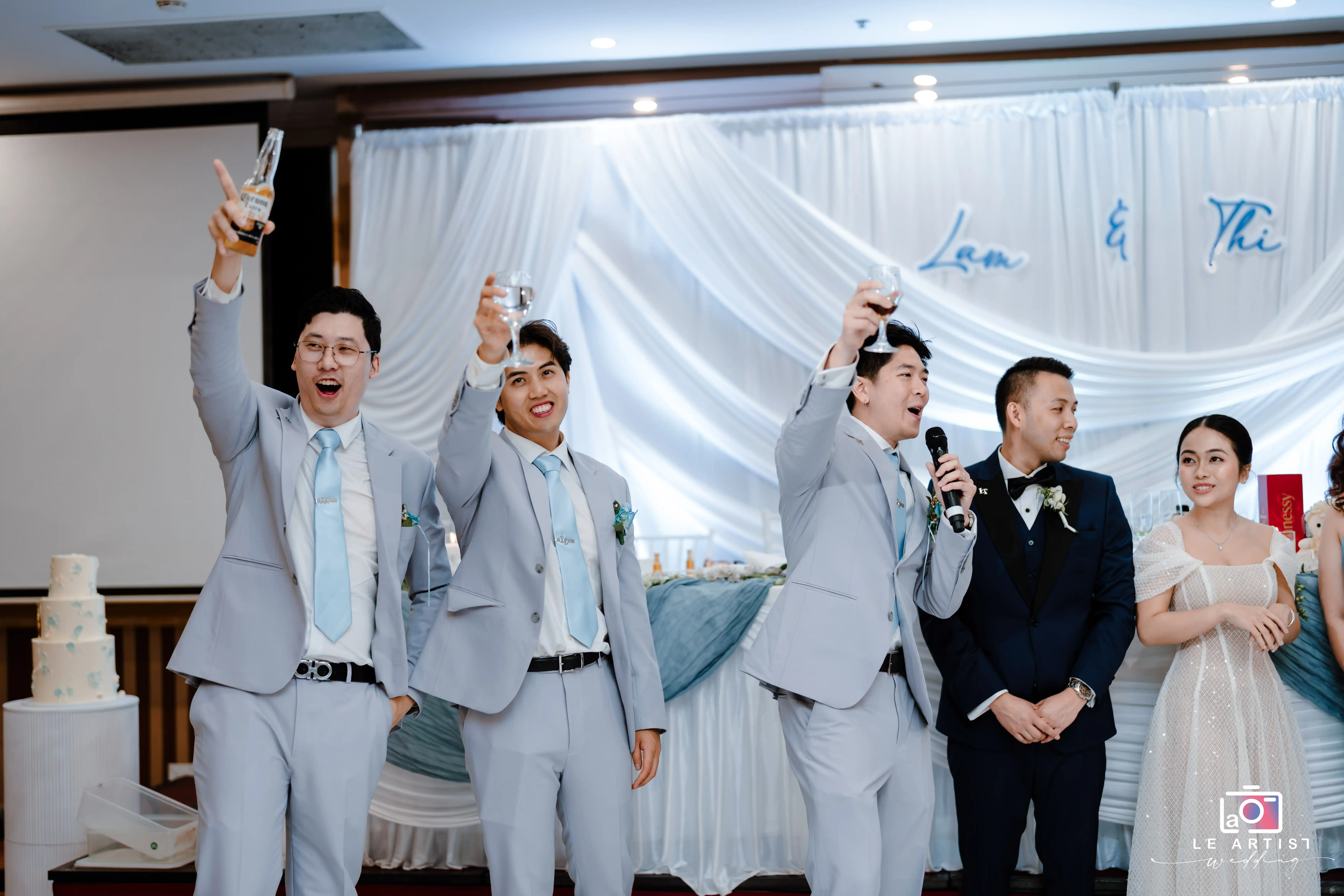 A lively scene from the wedding reception where wedding party members are enthusiastically raising their glasses in a toast. The backdrop features elegant drapery and the couple's names, adding to the festive atmosphere. Le Artist Wedding