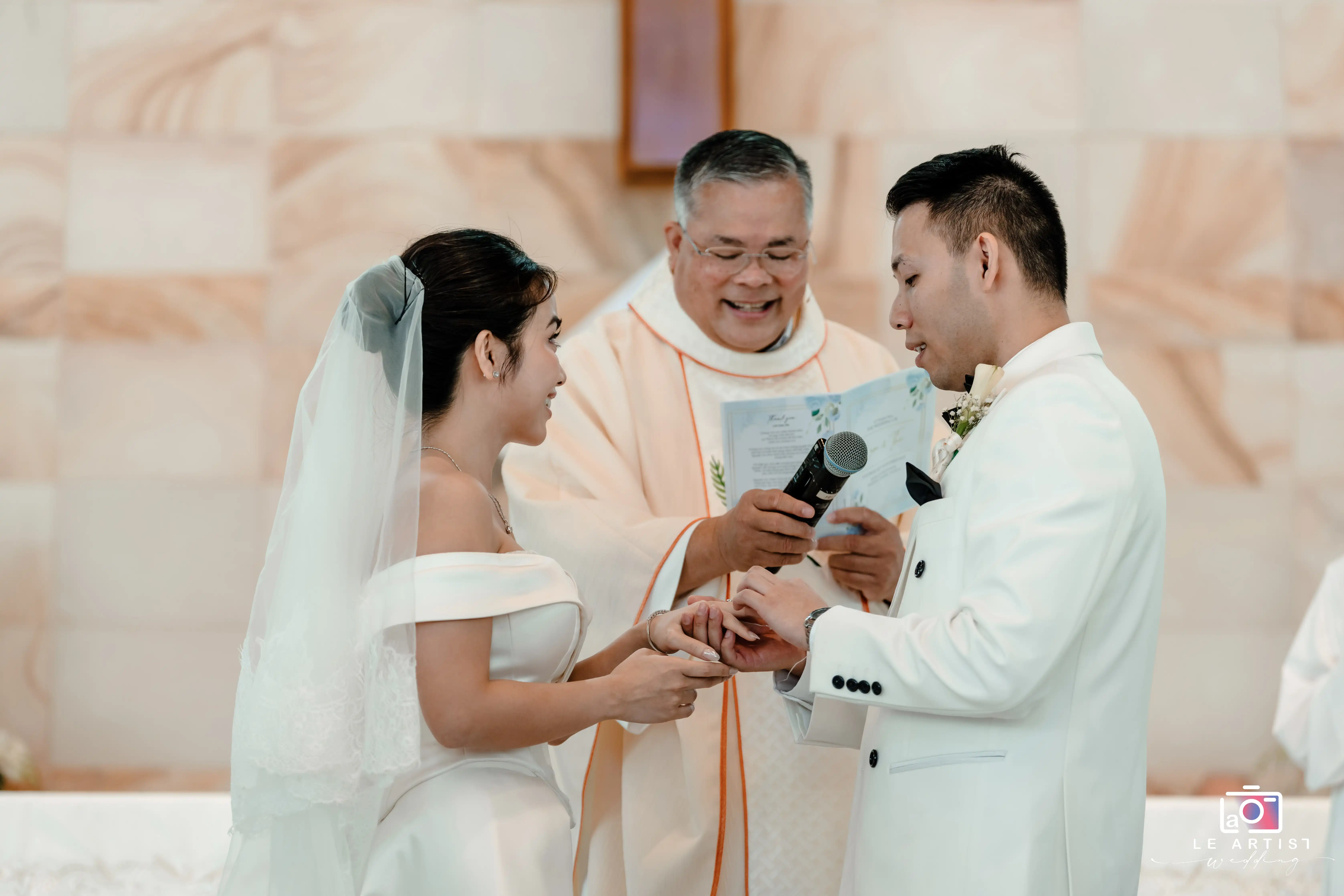 The couple exchanges rings during their wedding ceremony, sealing their vows of love and commitment in front of a joyful officiant. Le Artist Wedding