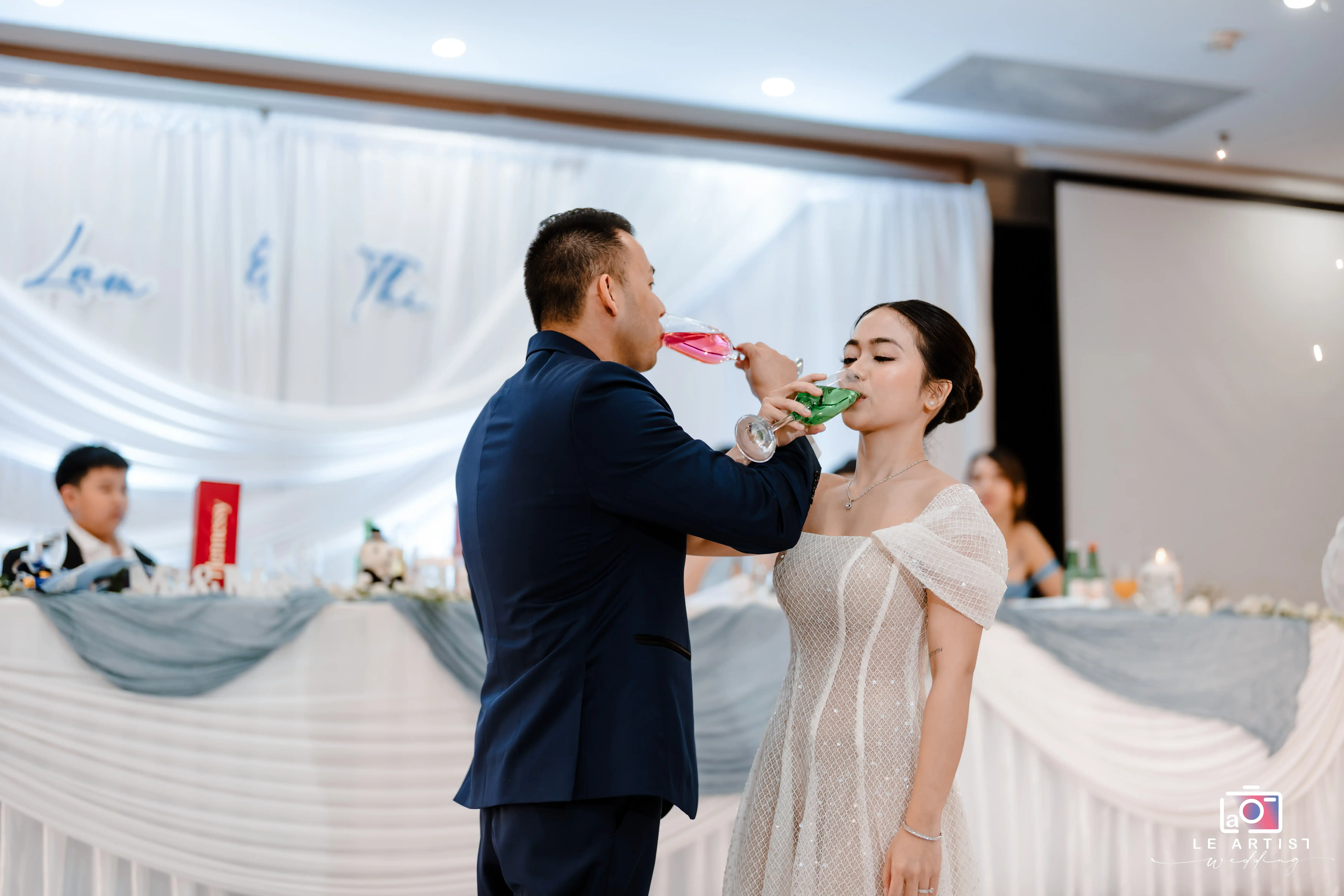 The couple shares a toast during their wedding reception, elegantly dressed and surrounded by a beautifully decorated backdrop. Le Artist Wedding