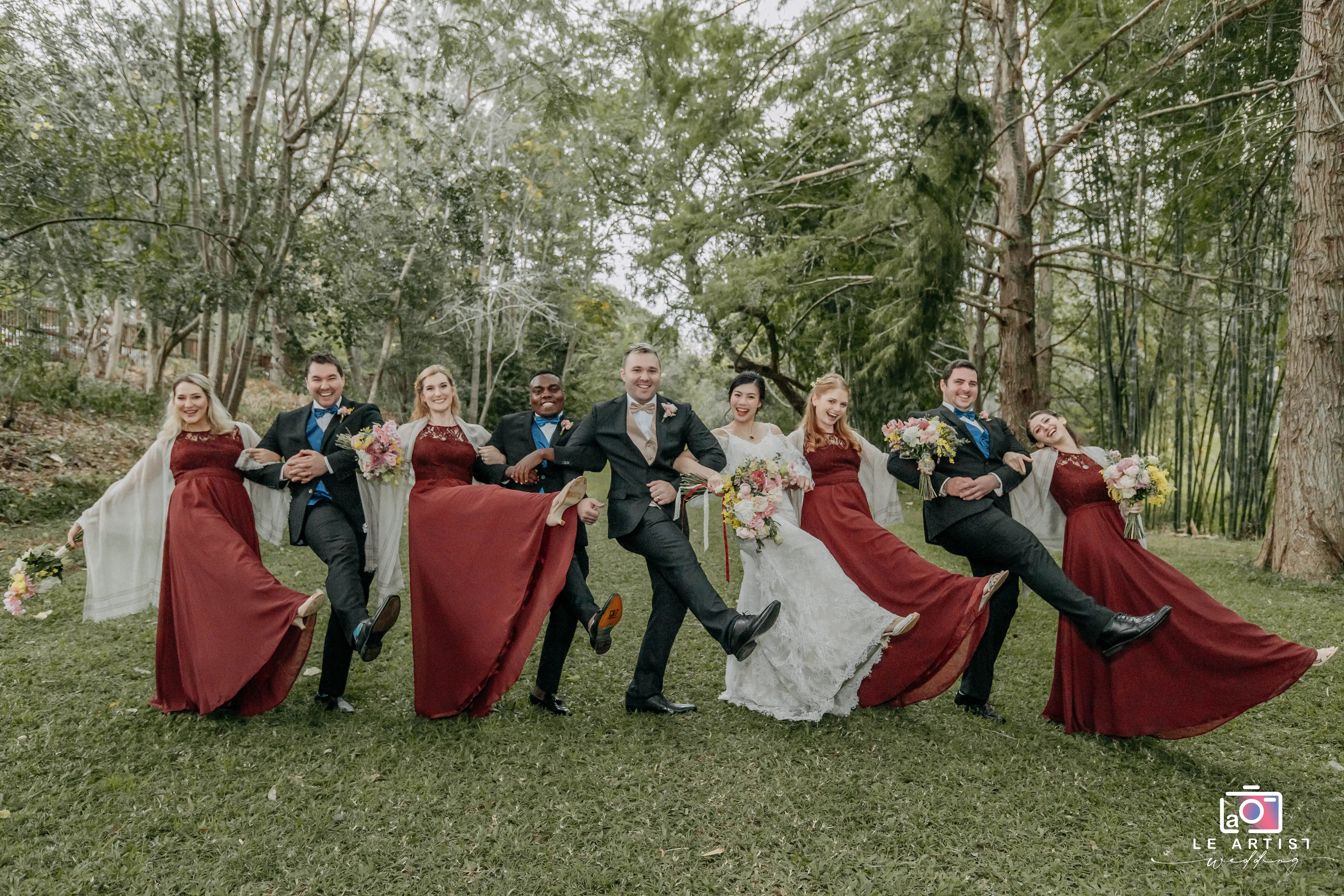 The bridal party performs a joyful kickline, showcasing their vibrant personalities and camaraderie, set against a lush, wooded backdrop. Le Artist Wedding
