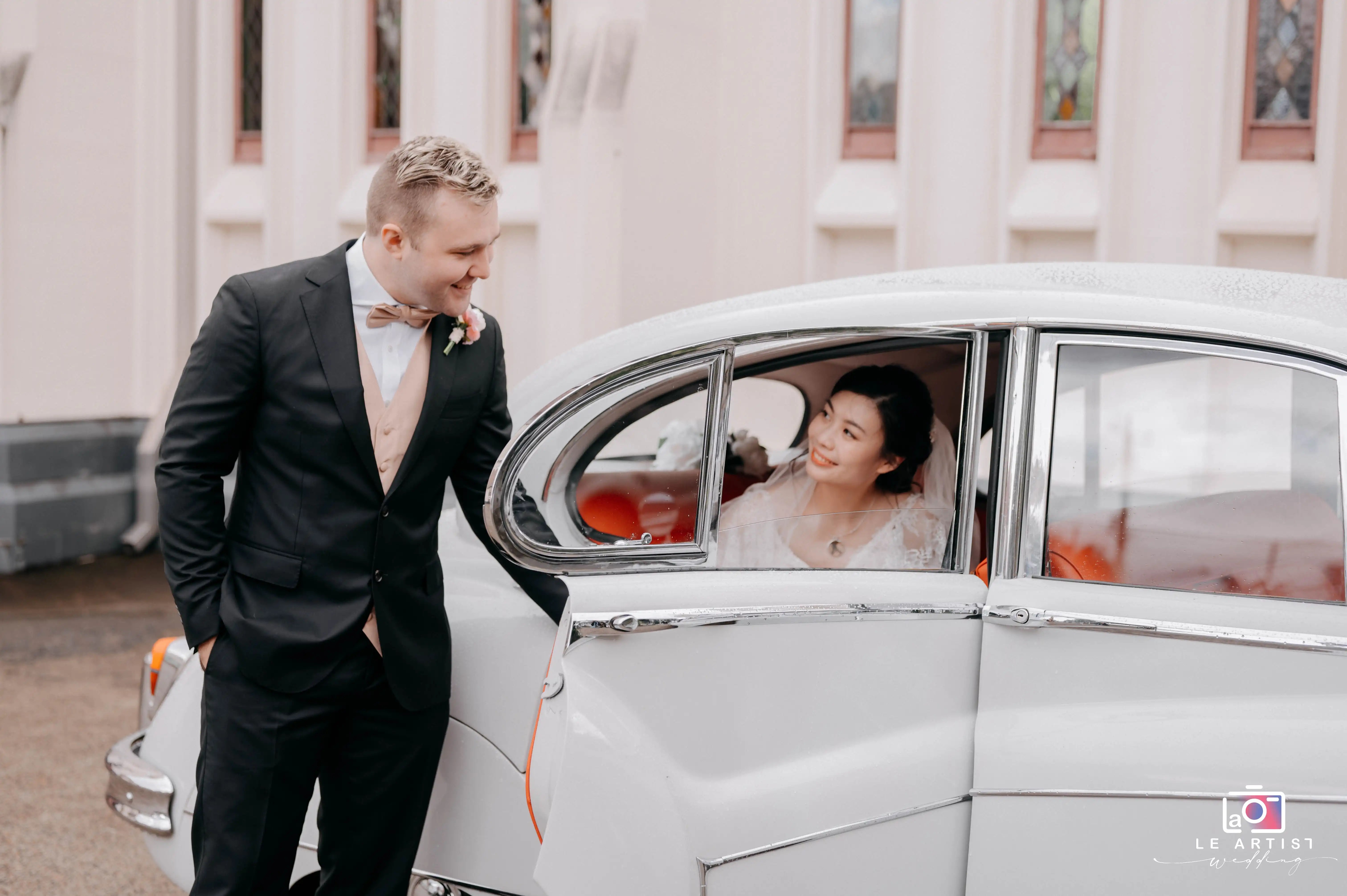A joyful moment as the groom leans toward the bride in the car, sharing a happy glance outside the church. Le Artist Wedding
