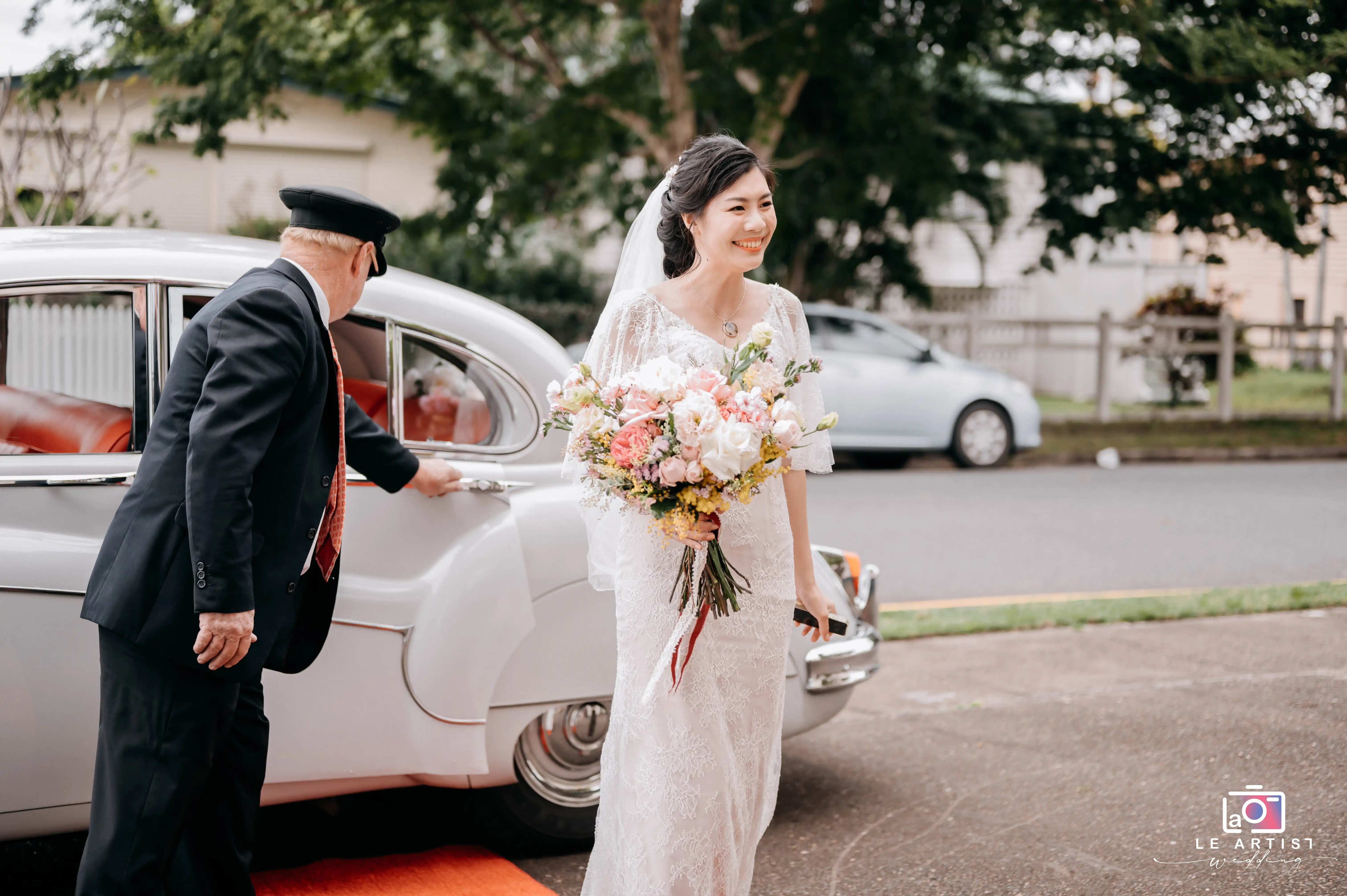 A joyful bride arrives, greeted by a chauffeur, holding a beautiful bouquet of pastel flowers, stepping out onto a red carpet. Le Artist Wedding