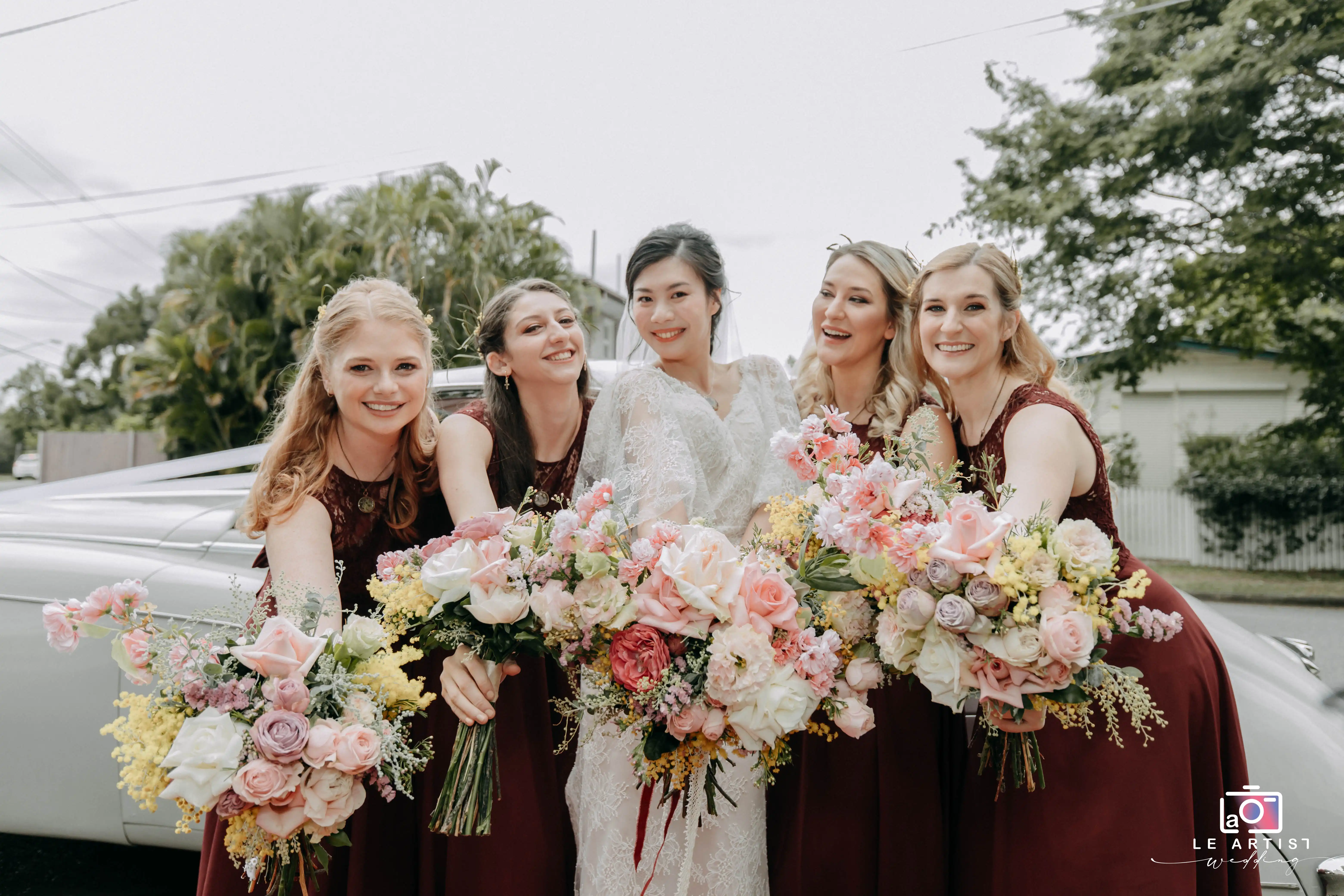 The bride and her bridesmaids joyfully pose with their colorful bouquets in front of a classic car, exuding elegance and happiness. Le Artist Wedding