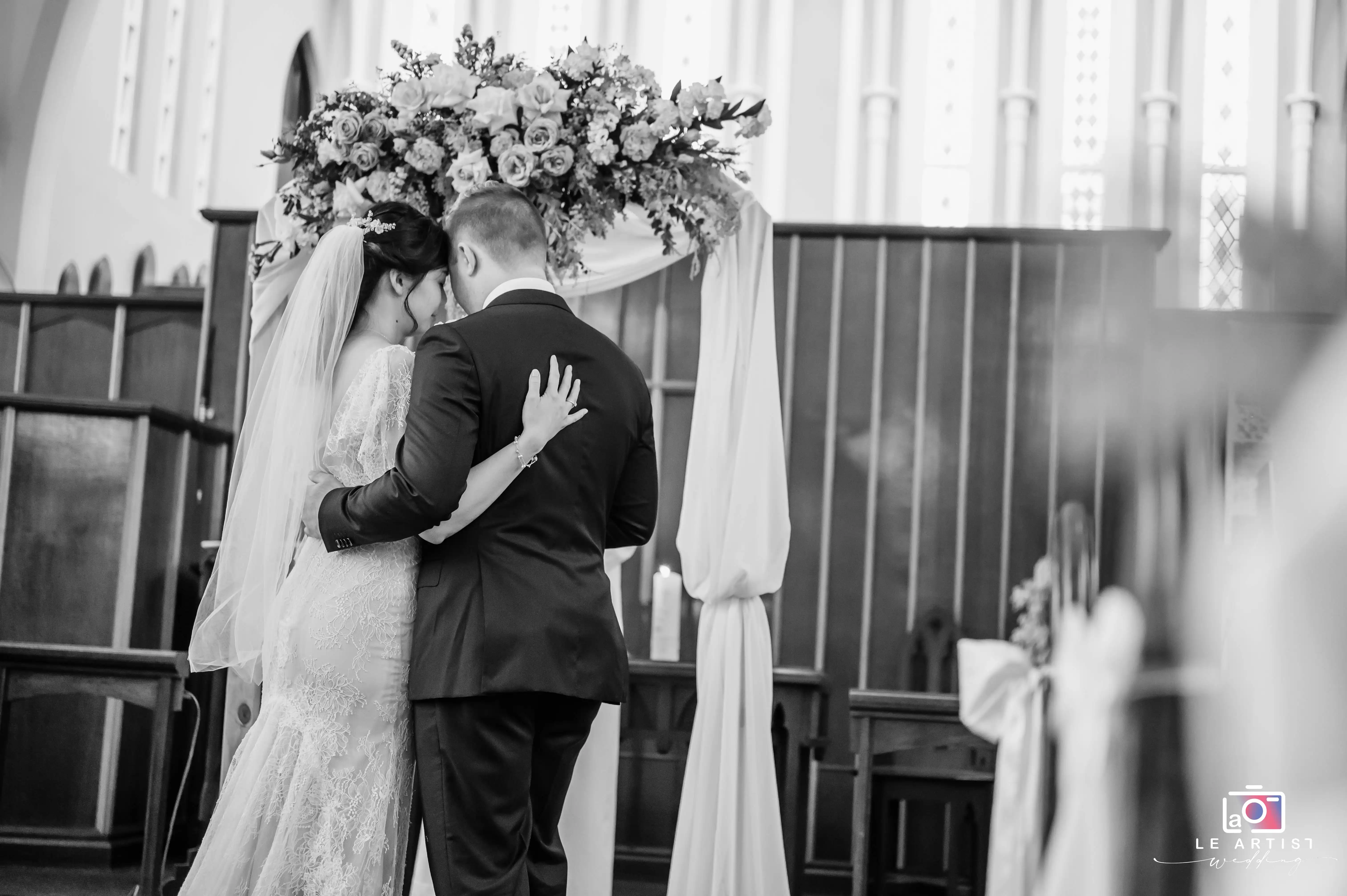 A tender moment captured as the newlyweds embrace beneath a beautifully adorned floral arch inside a church, symbolizing the beginning of their journey together. Le Artist Wedding