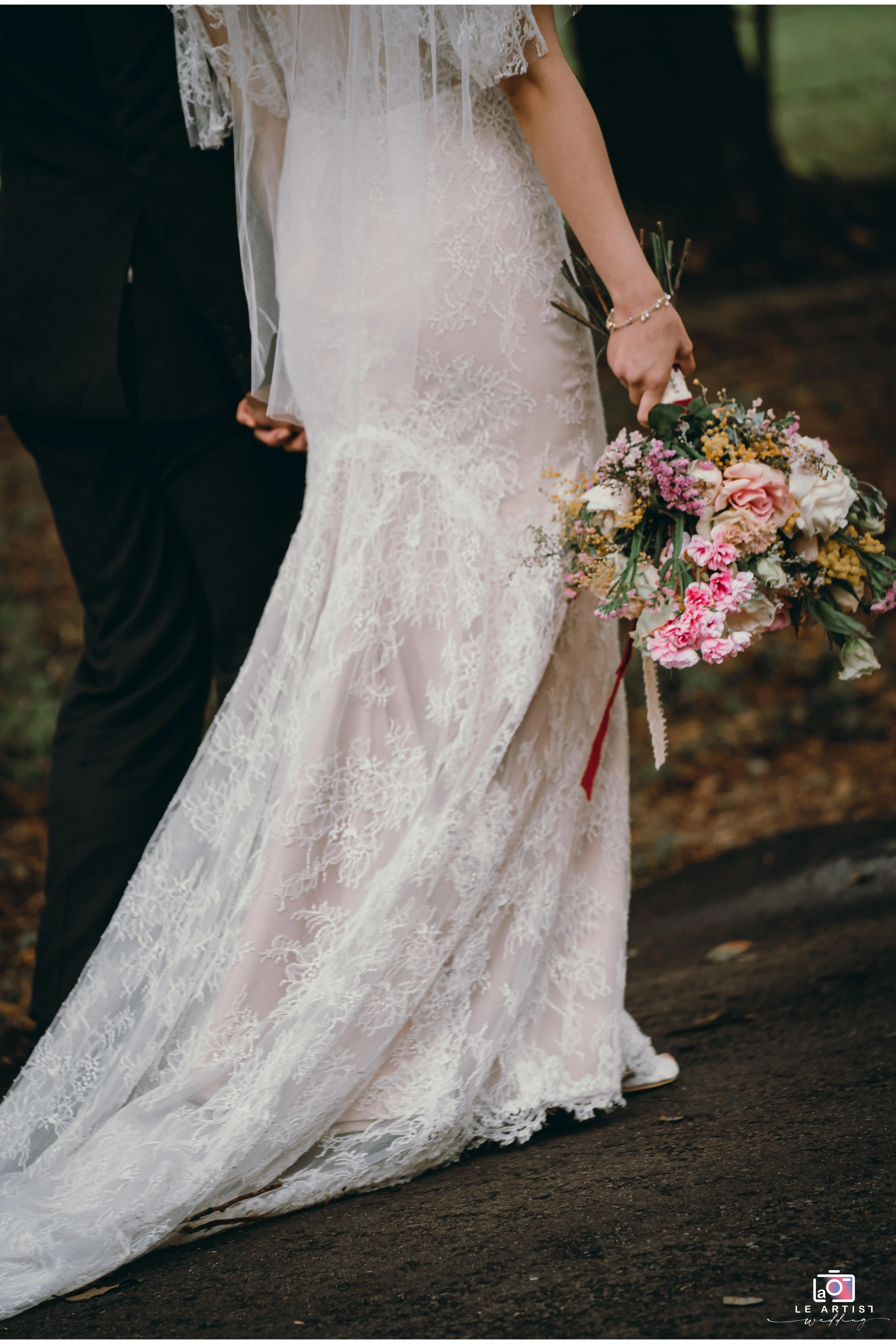The newlyweds walk hand in hand, capturing a beautiful moment of togetherness. The bride's lace gown and bouquet of vibrant flowers add elegance to this intimate scene. Le Artist Wedding