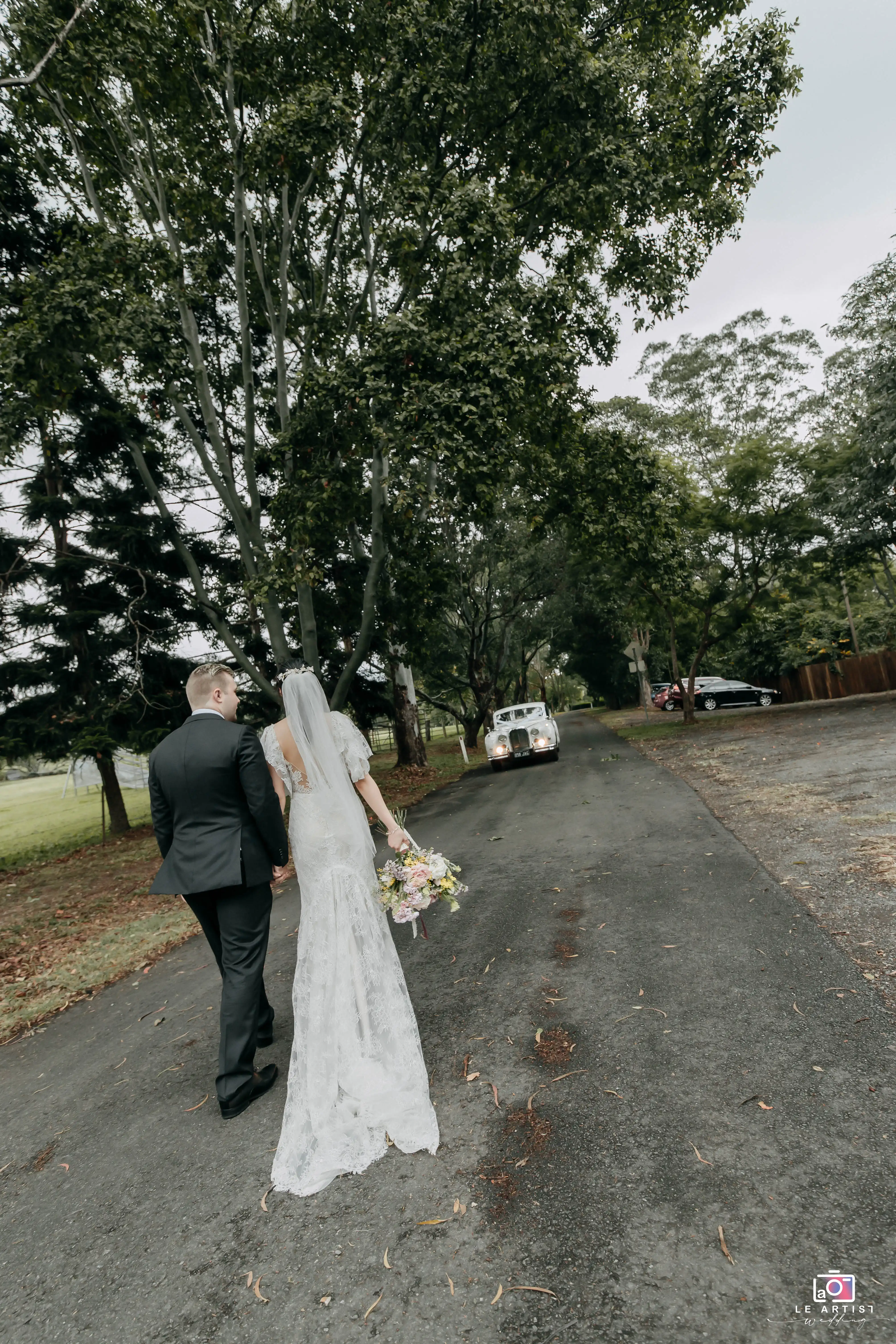 The newlyweds walk hand in hand along a tree-lined path, with a classic car waiting ahead. Le Artist Wedding