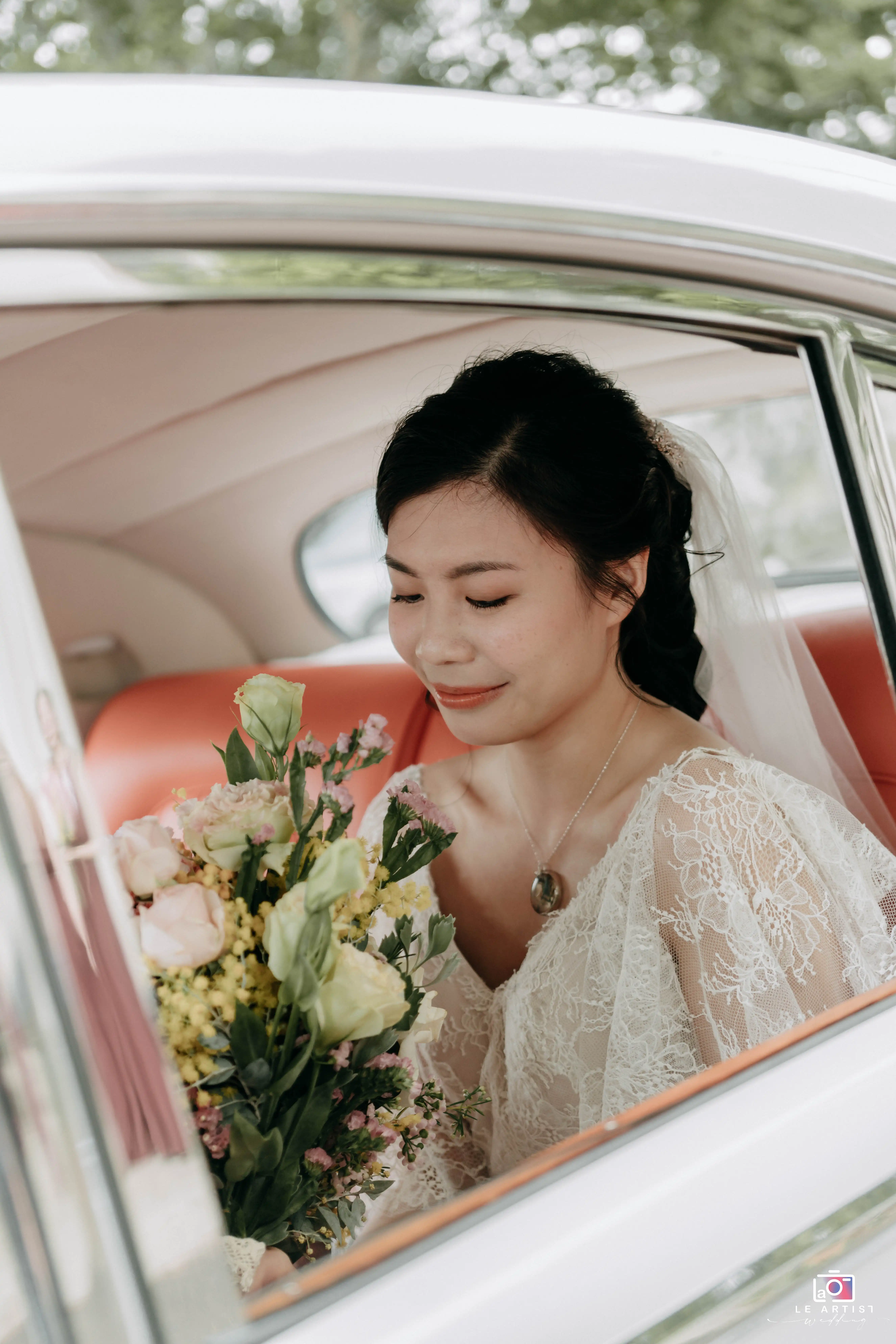 A bride sits gracefully in a car, holding her bouquet and wearing a delicate lace dress with a serene smile. Le Artist Wedding
