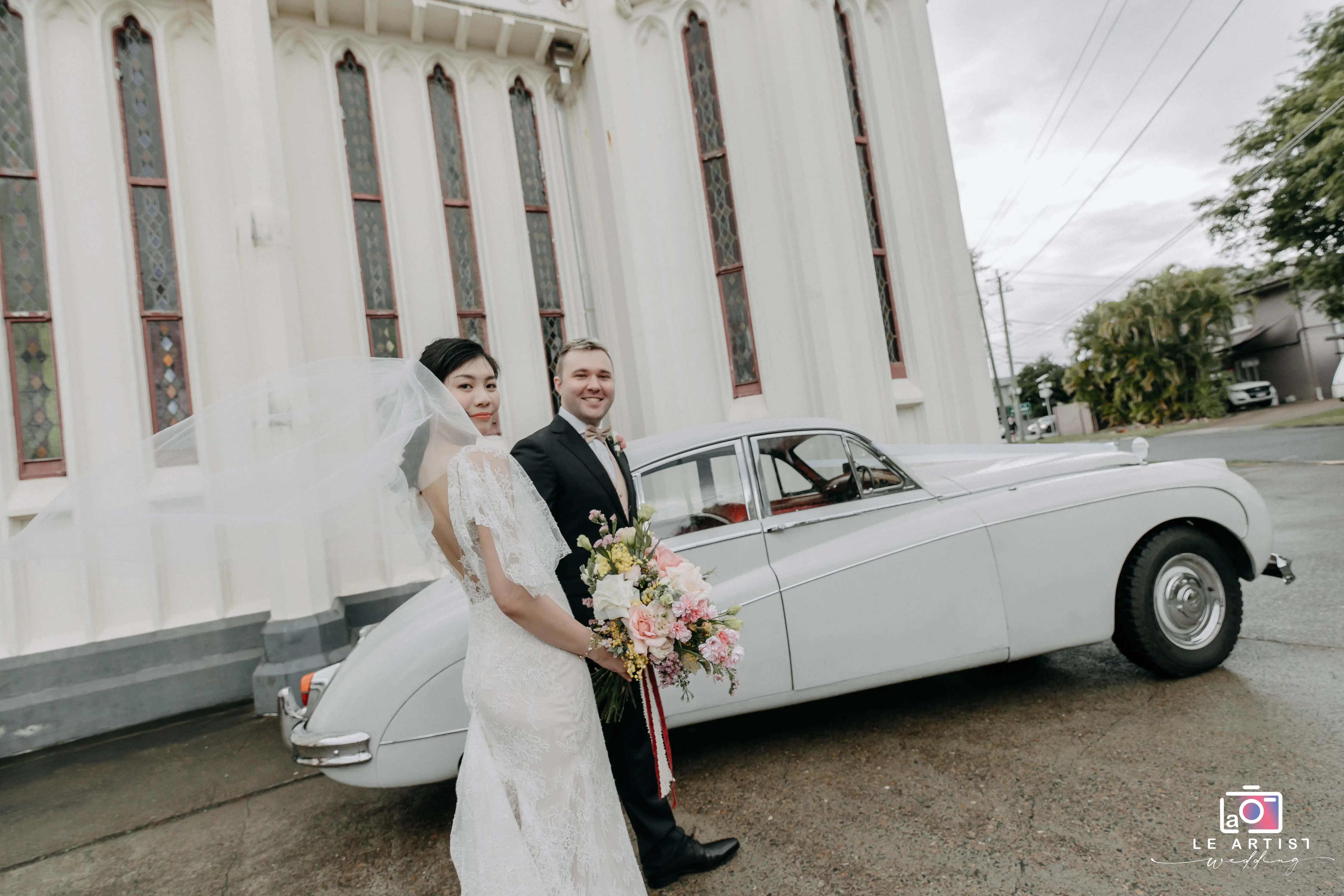 The newlyweds pose elegantly in front of a classic car beside a historic church, capturing a timeless moment with bouquets and smiles. Le Artist Wedding