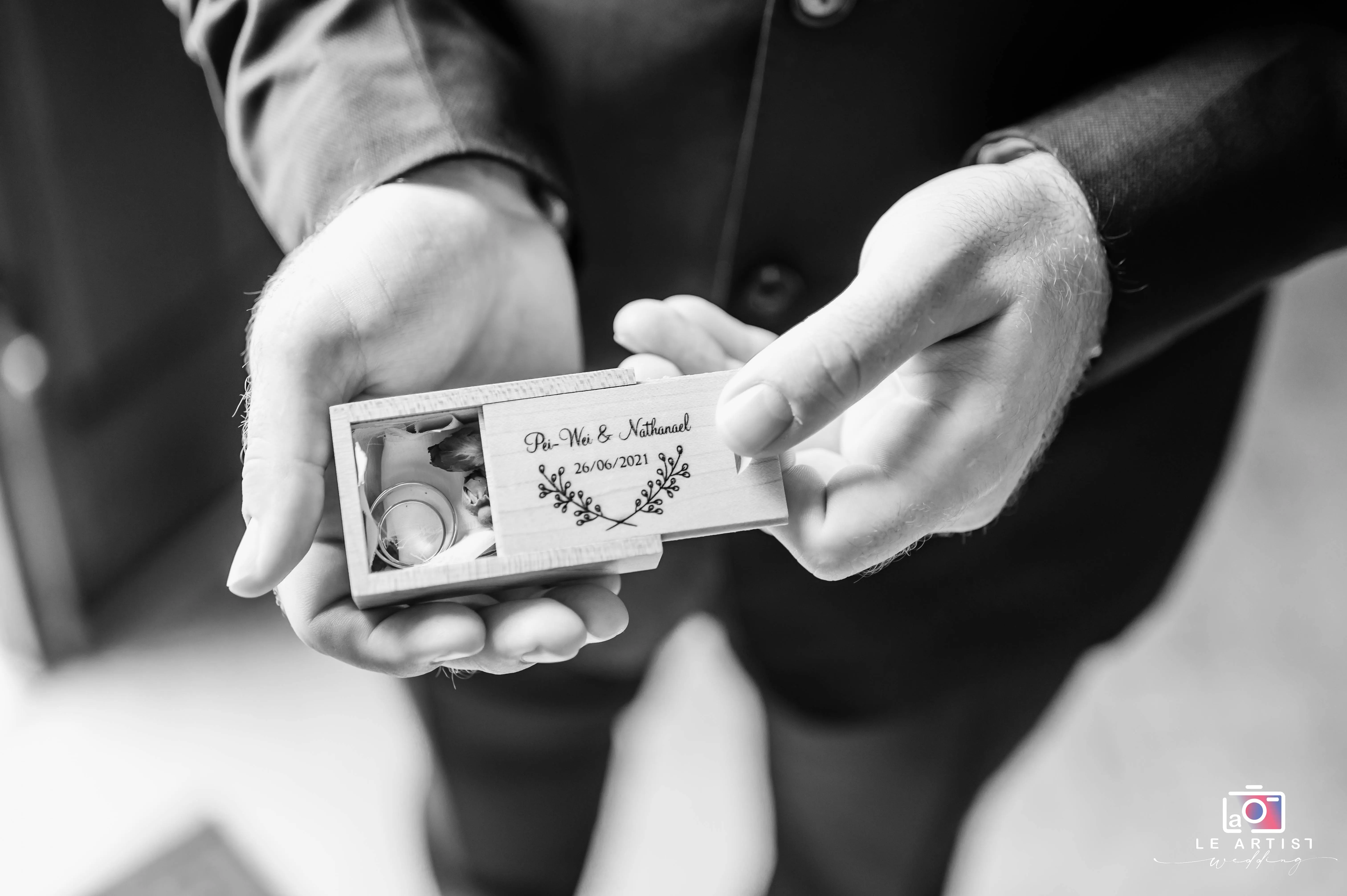 A close-up of a hands holding a personalized wooden ring box, elegantly engraved with the couple's names and wedding date. Le Artist Wedding