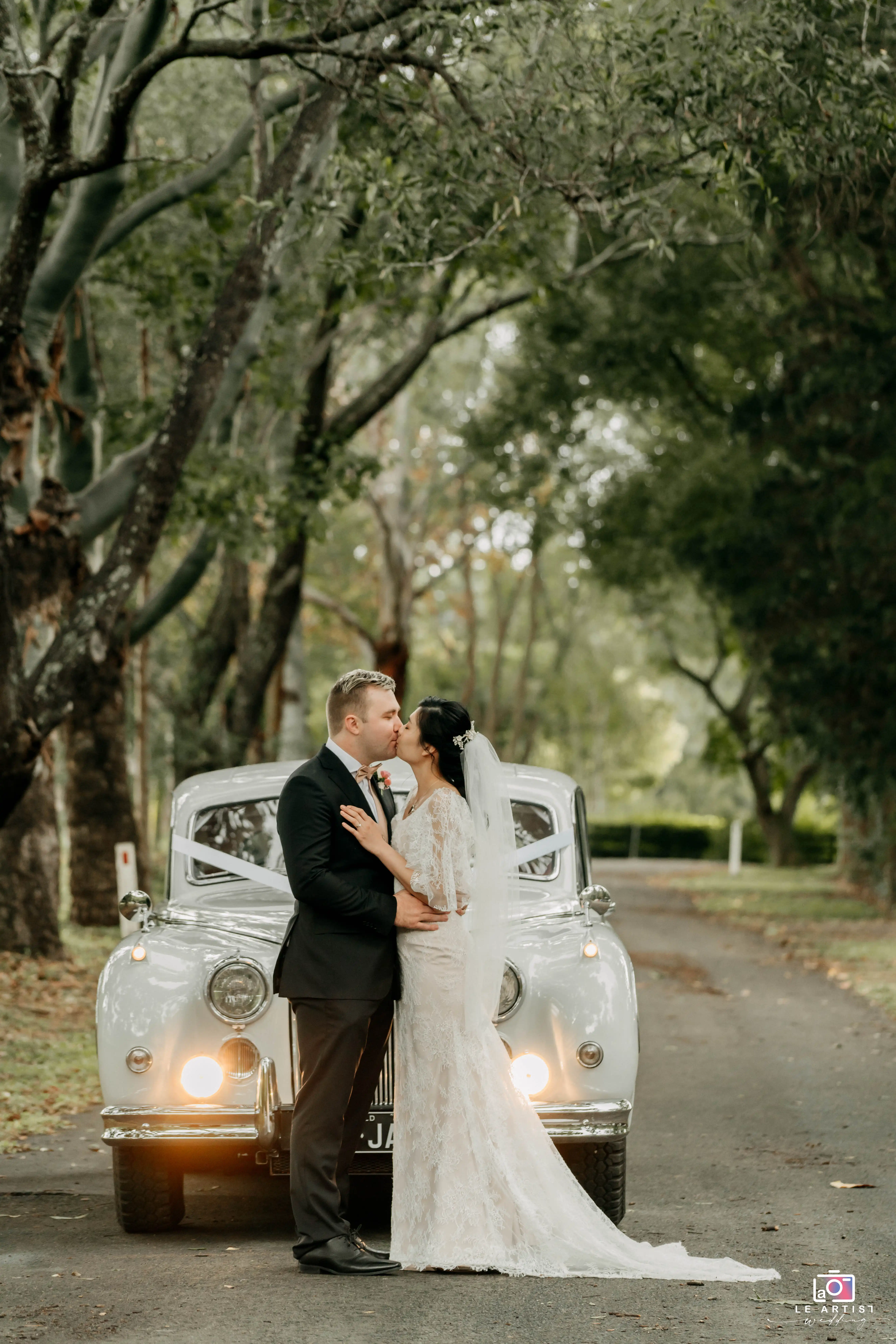 A newlywed couple shares a tender kiss in front of a vintage car lined with soft ribbons, surrounded by lush trees on a serene path. Le Artist Wedding