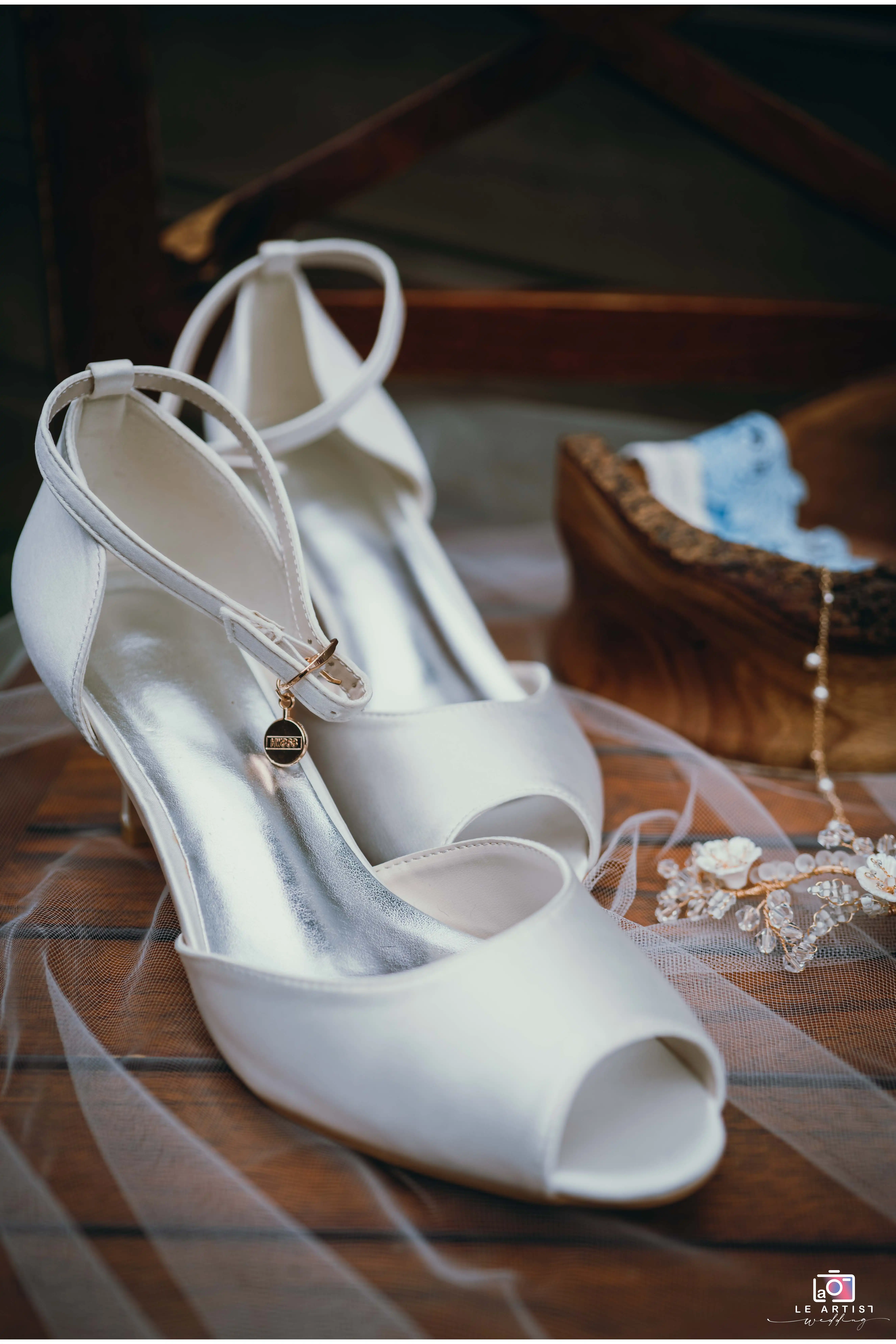 Elegant bridal shoes paired with delicate jewelry and a lace garter, captured on a wooden chair, setting a romantic tone before the wedding ceremony. Le Artist Wedding
