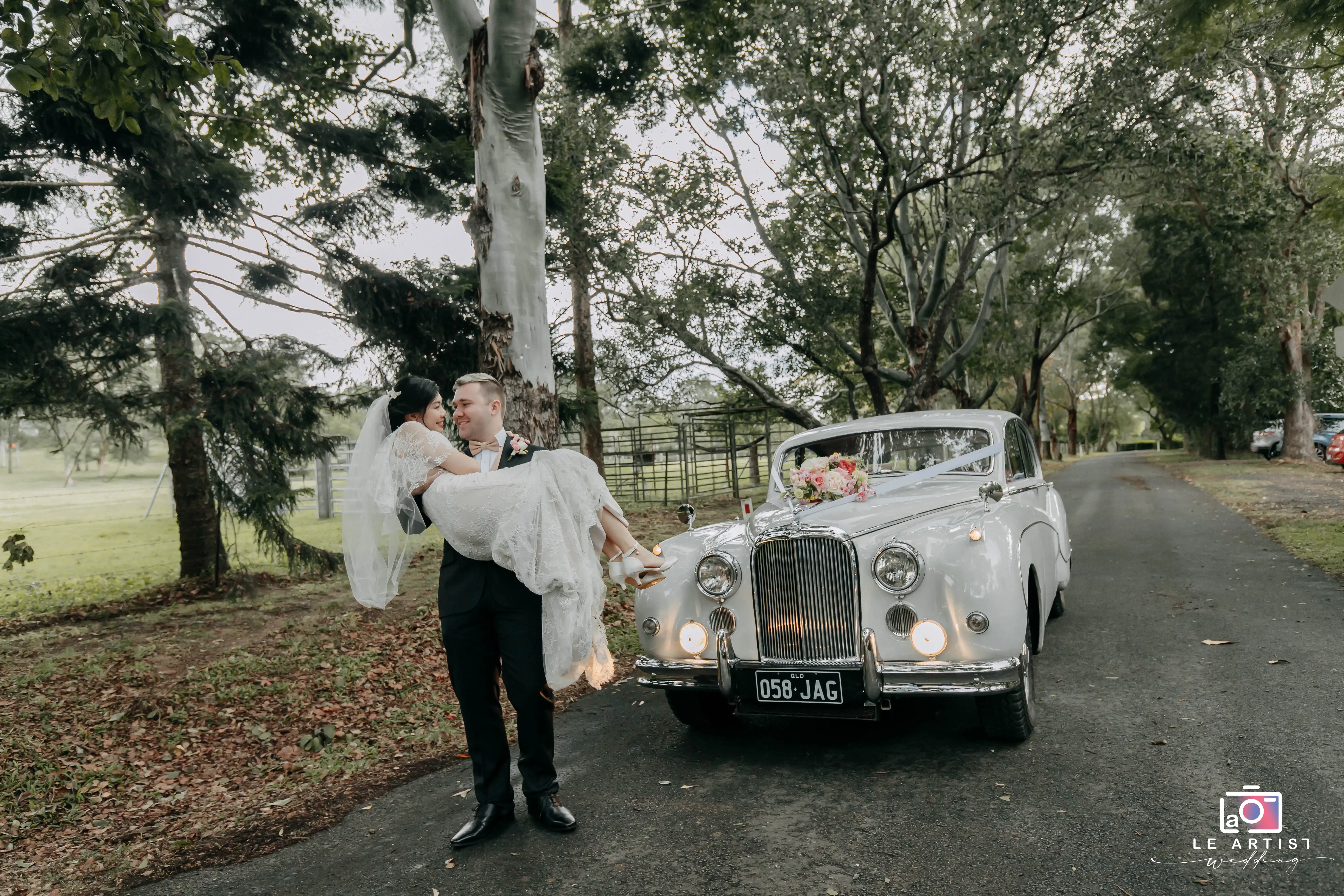 The groom carries his bride in front of a vintage car adorned with flowers, set against a serene, wooded backdrop. Le Artist Wedding