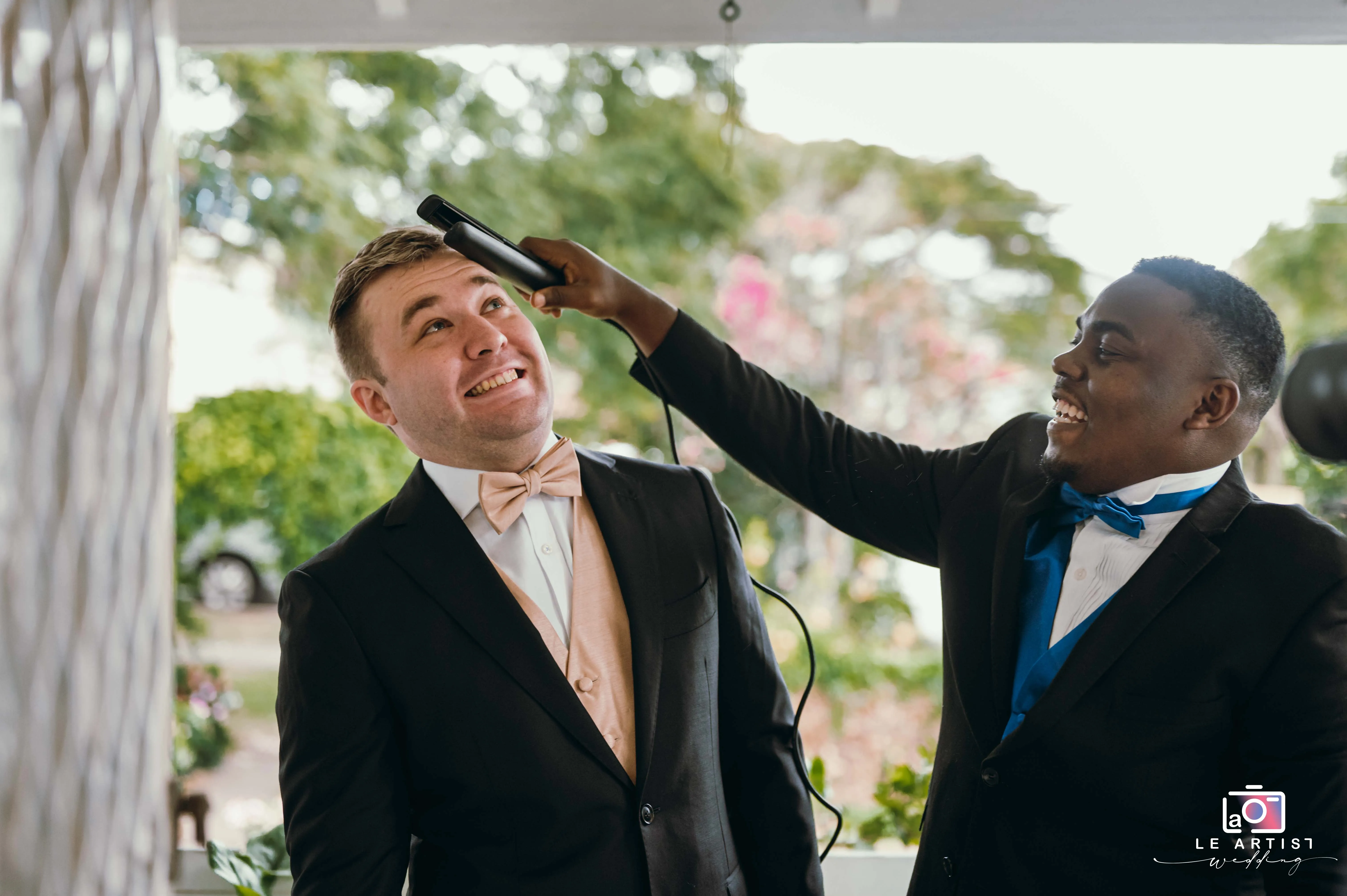 A joyful moment as the groom shares a laugh while having his hair styled, highlighting the camaraderie and fun during wedding preparations. Le Artist Wedding
