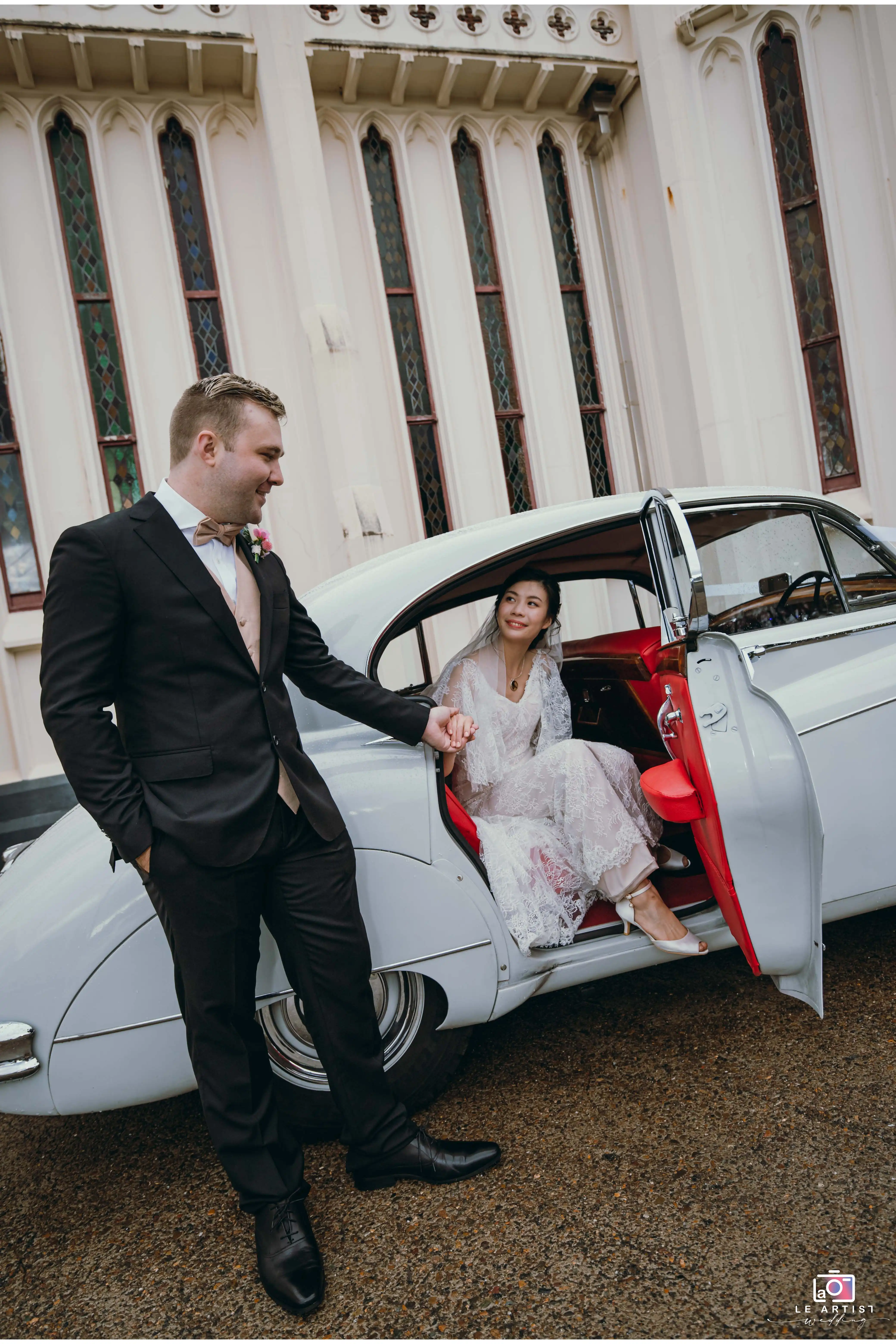 The groom lovingly helps the bride step out of a classic car, setting the scene for a beautiful wedding day. Le Artist Wedding