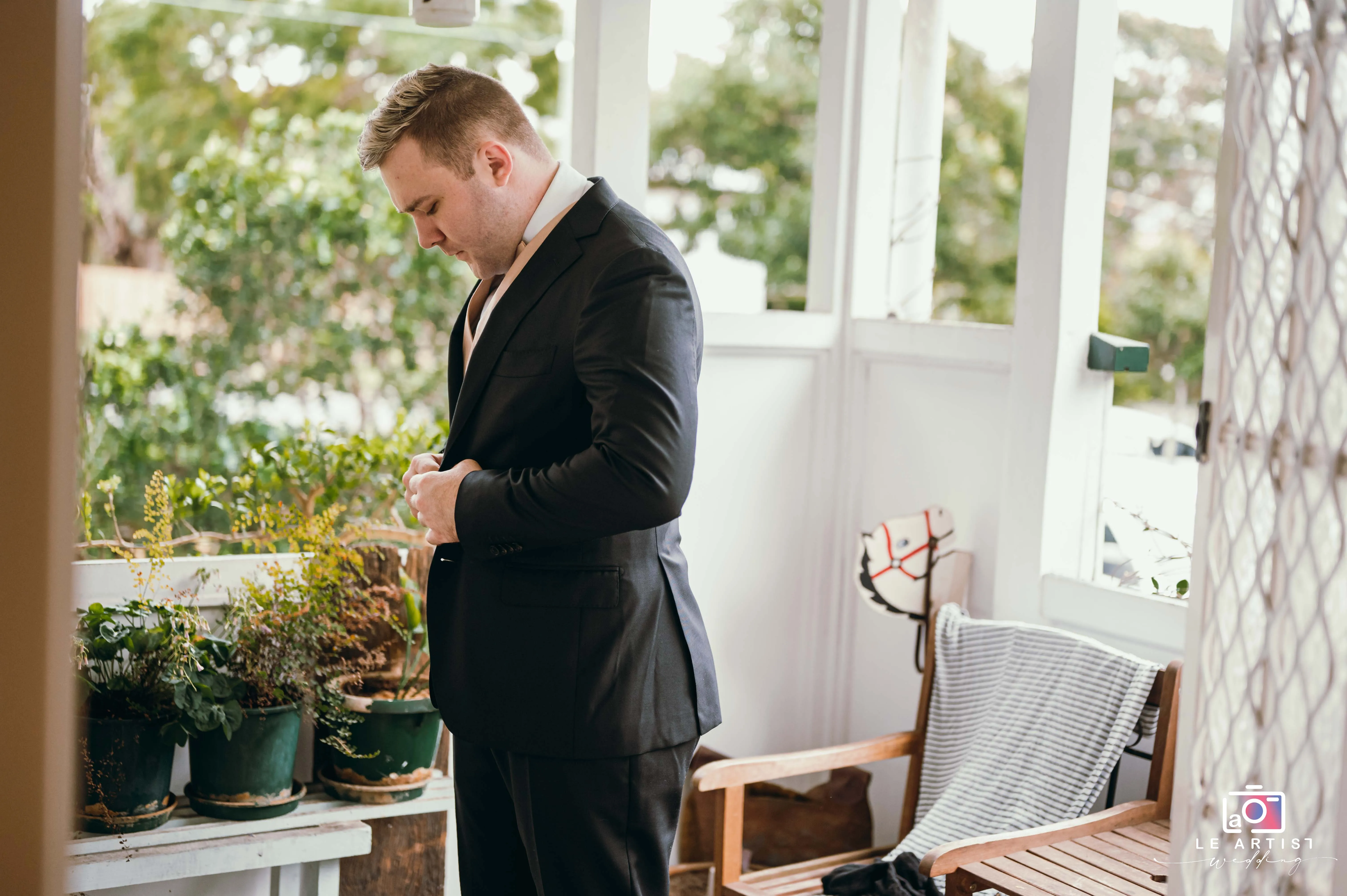 A groom meticulously buttoning his suit jacket, preparing for the wedding day ahead, surrounded by a serene outdoor setting. Le Artist Wedding