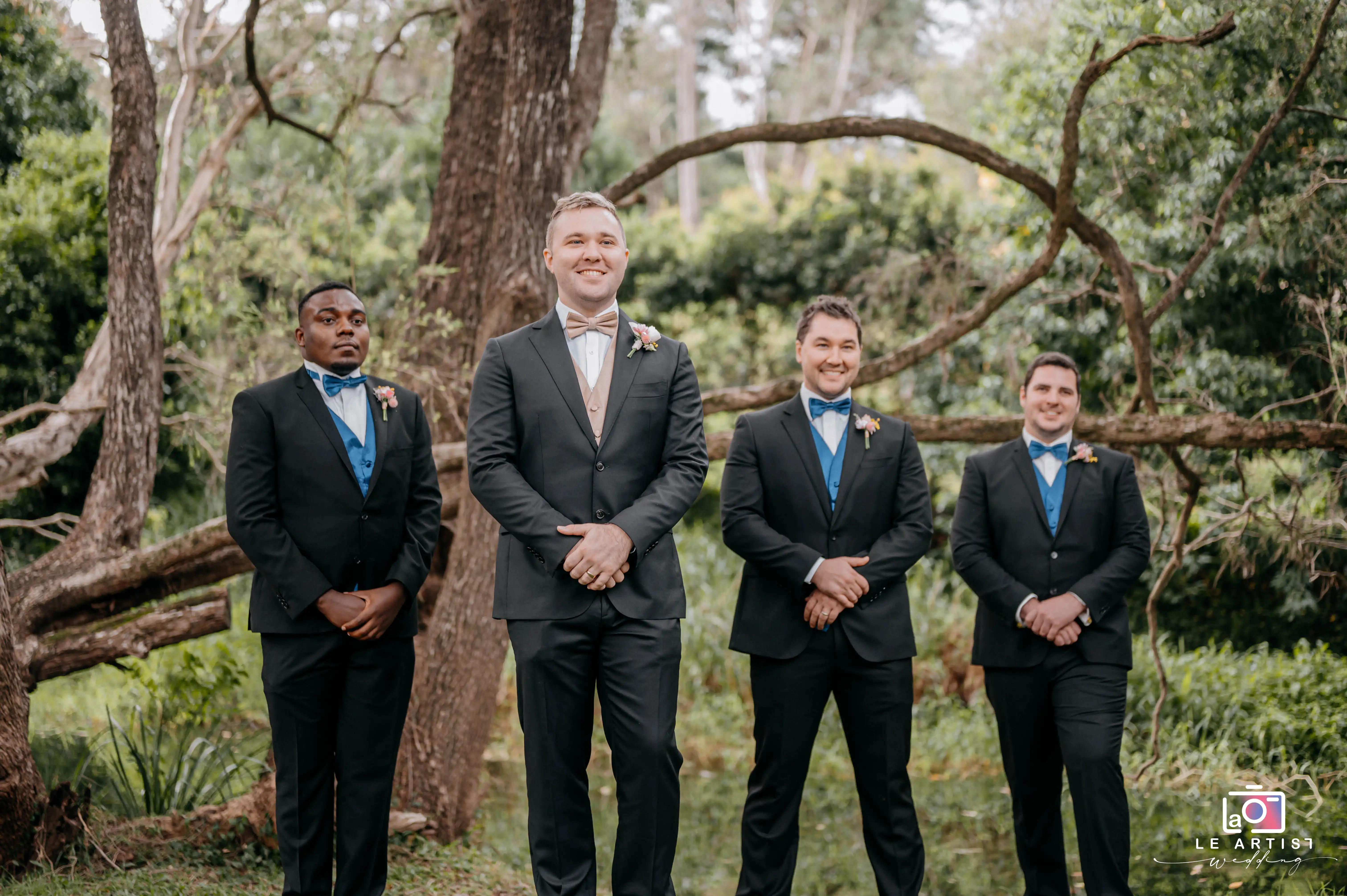 The groom and his groomsmen stand proudly in a lush, wooded setting, showcasing their formal attire and boutonnieres, ready for the ceremony. Le Artist Wedding
