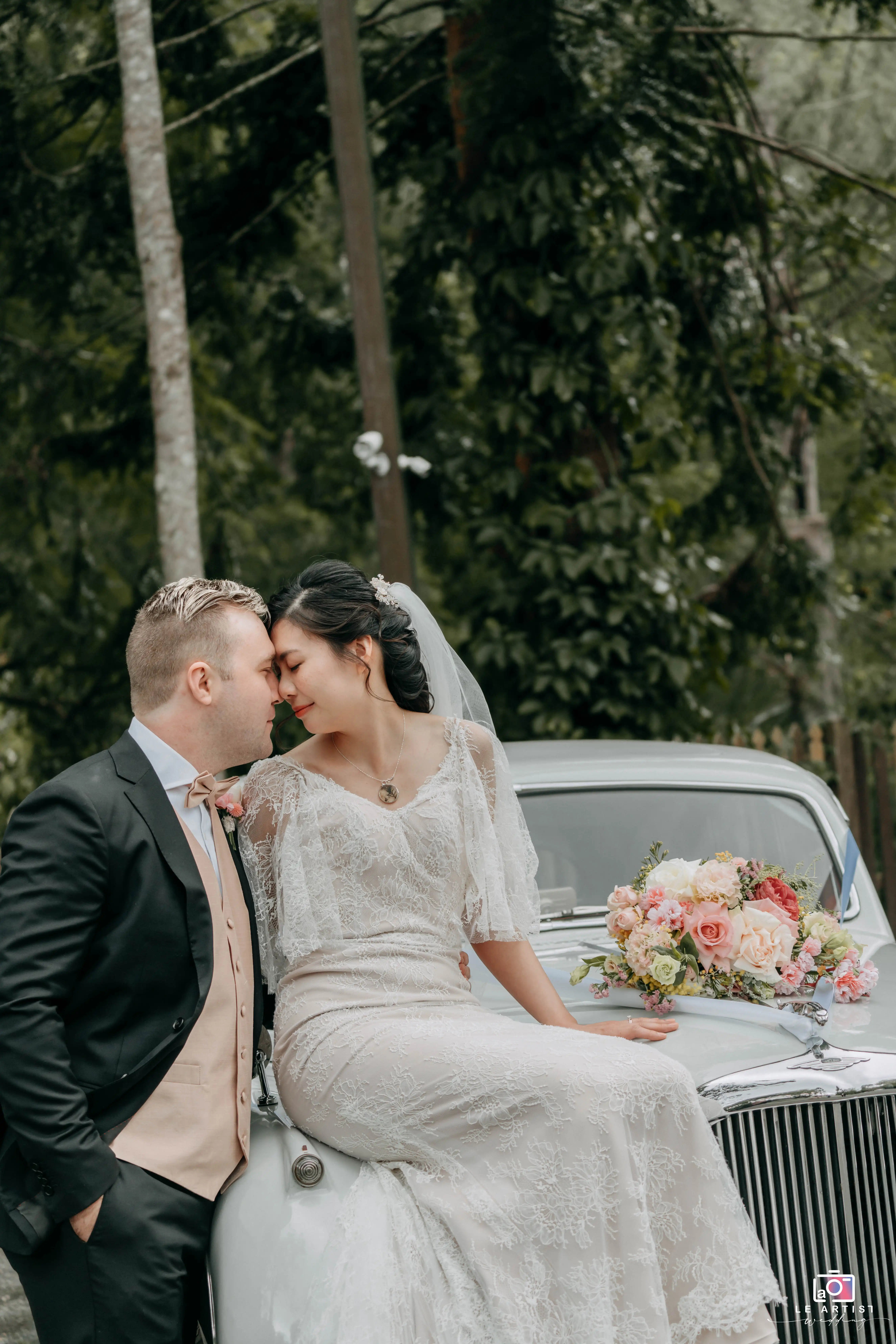 The bride and groom share an intimate moment on the classic car's hood, surrounded by the lush greenery, with a bouquet of flowers adding charm to the scene. Le Artist Wedding