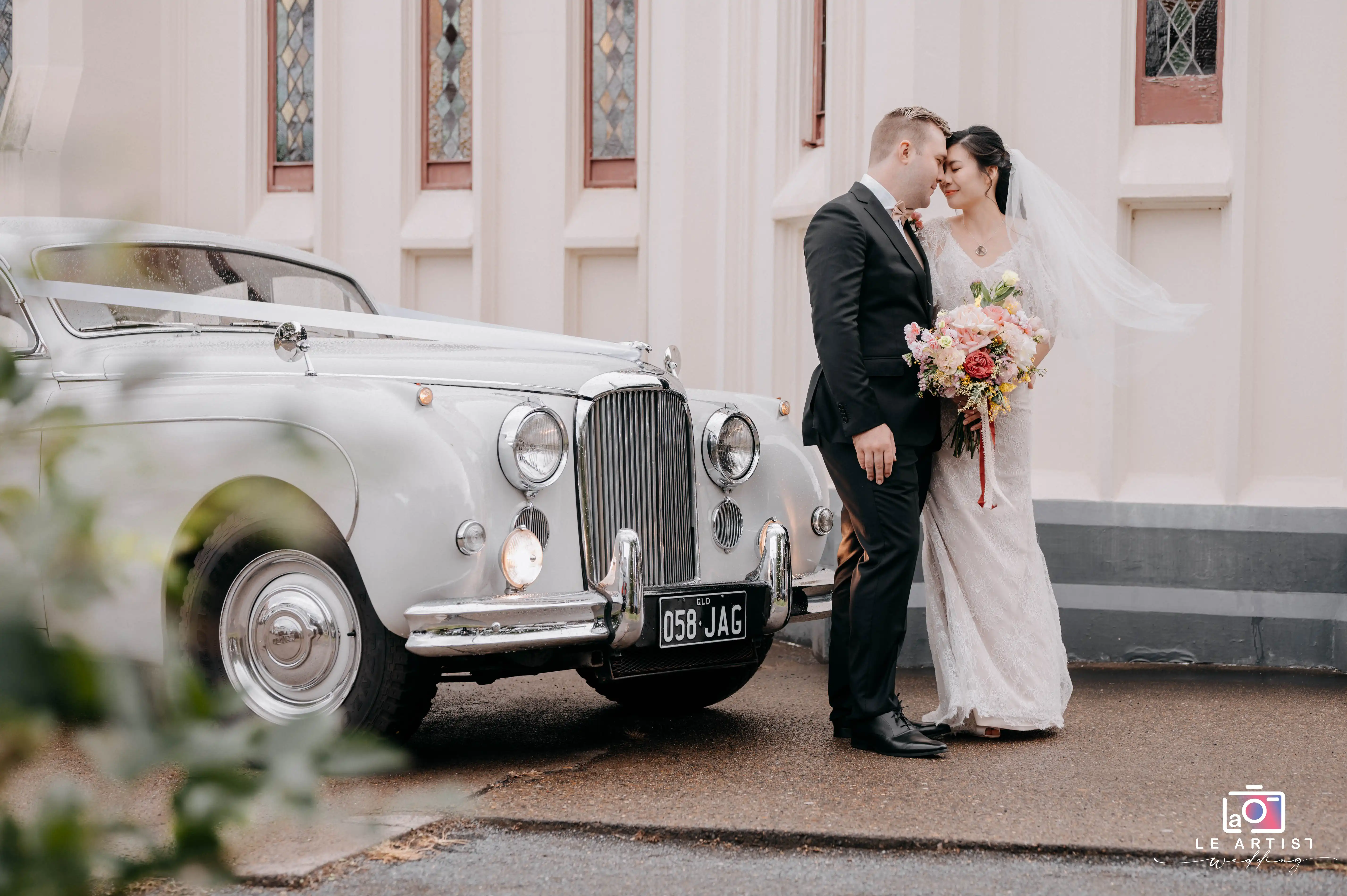 A newlywed couple shares a tender moment by a classic car, with the bride holding a colorful bouquet. The background features elegant architecture, adding to the charm of the scene. Le Artist Wedding