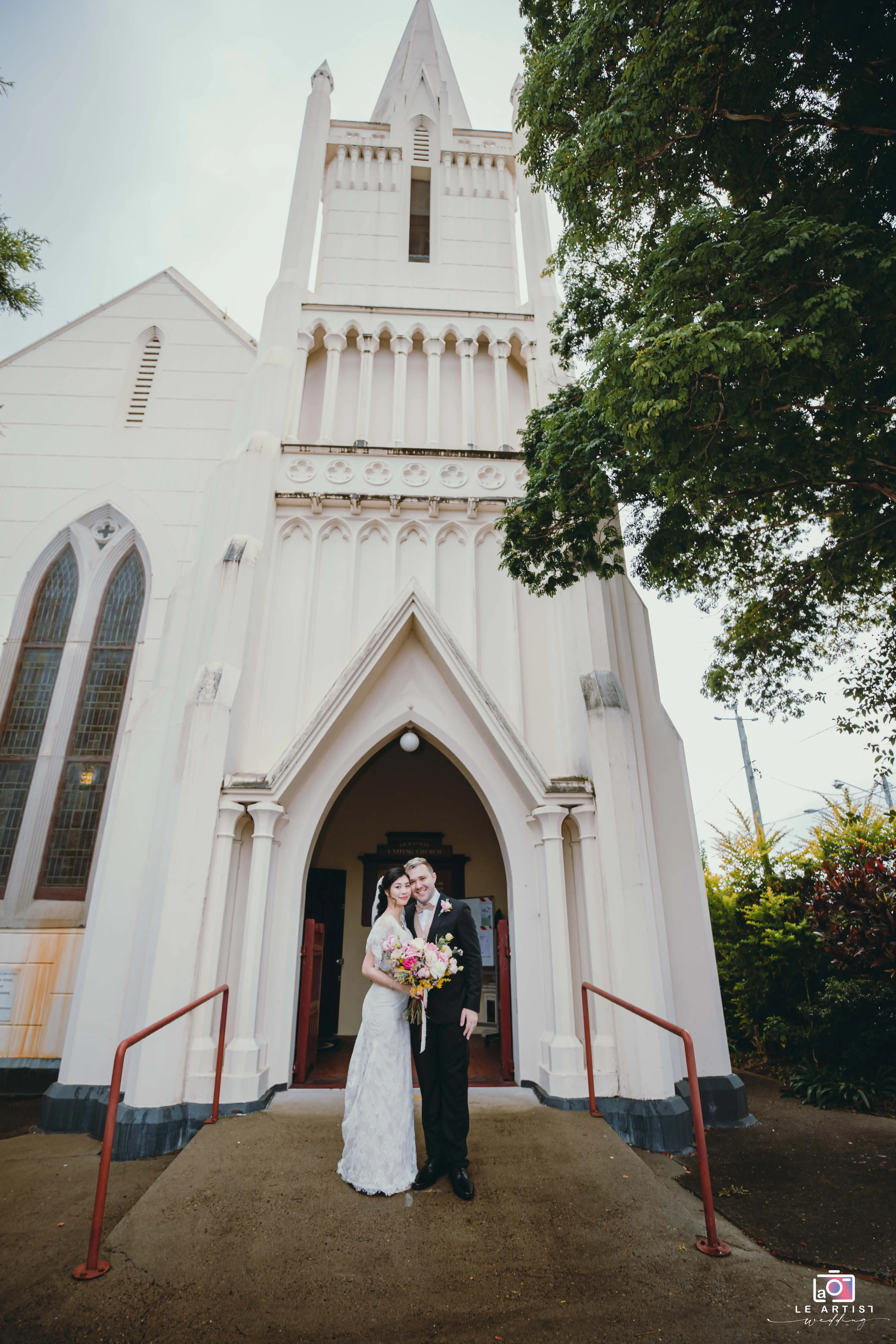 The newlywed couple stands happily in front of a grand church entrance, holding a vibrant bouquet of flowers, capturing a perfect post-ceremony moment. Le Artist Wedding