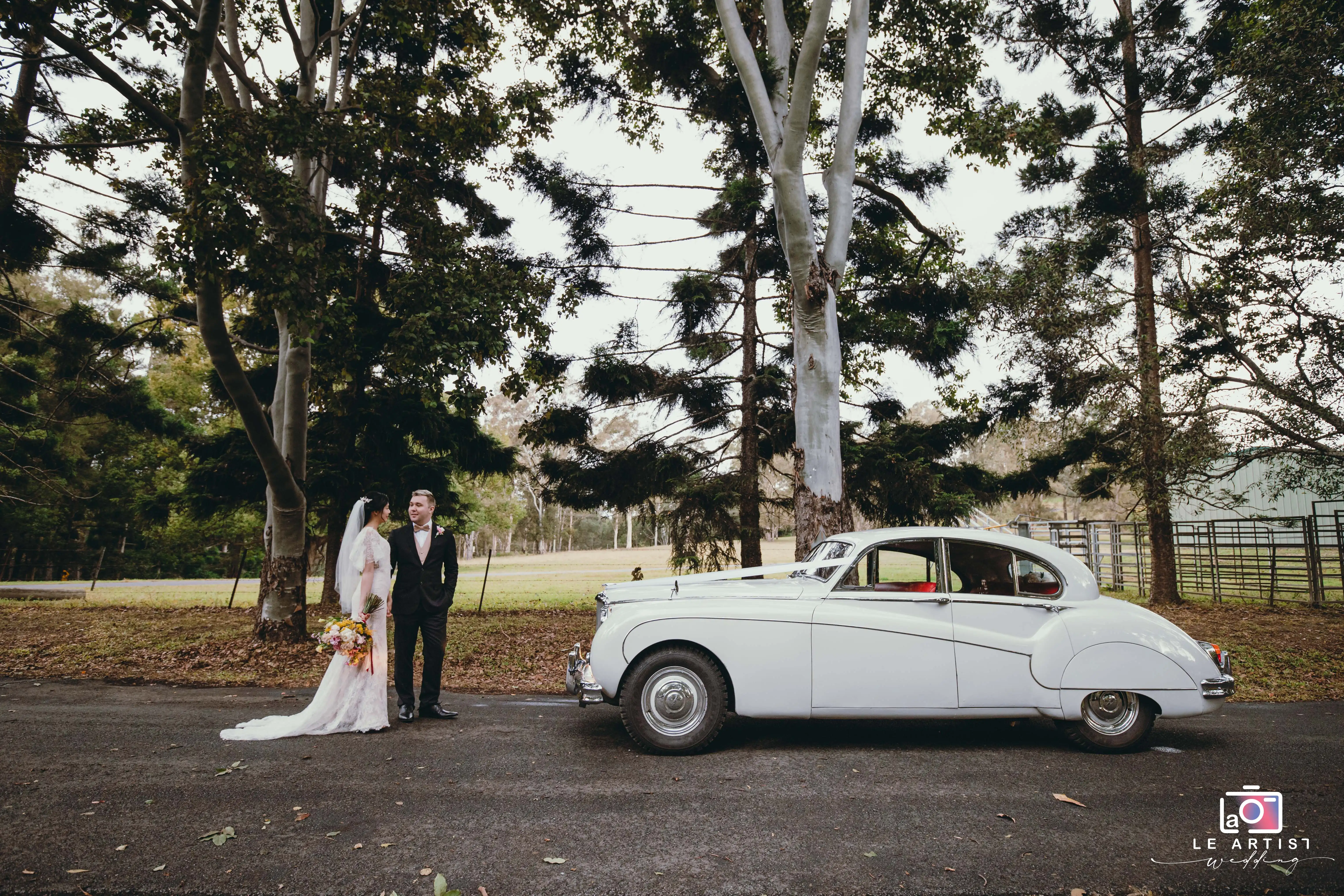 The newlyweds share a moment beside a classic white car, framed by towering trees, capturing their joy and style on their special day. Le Artist Wedding