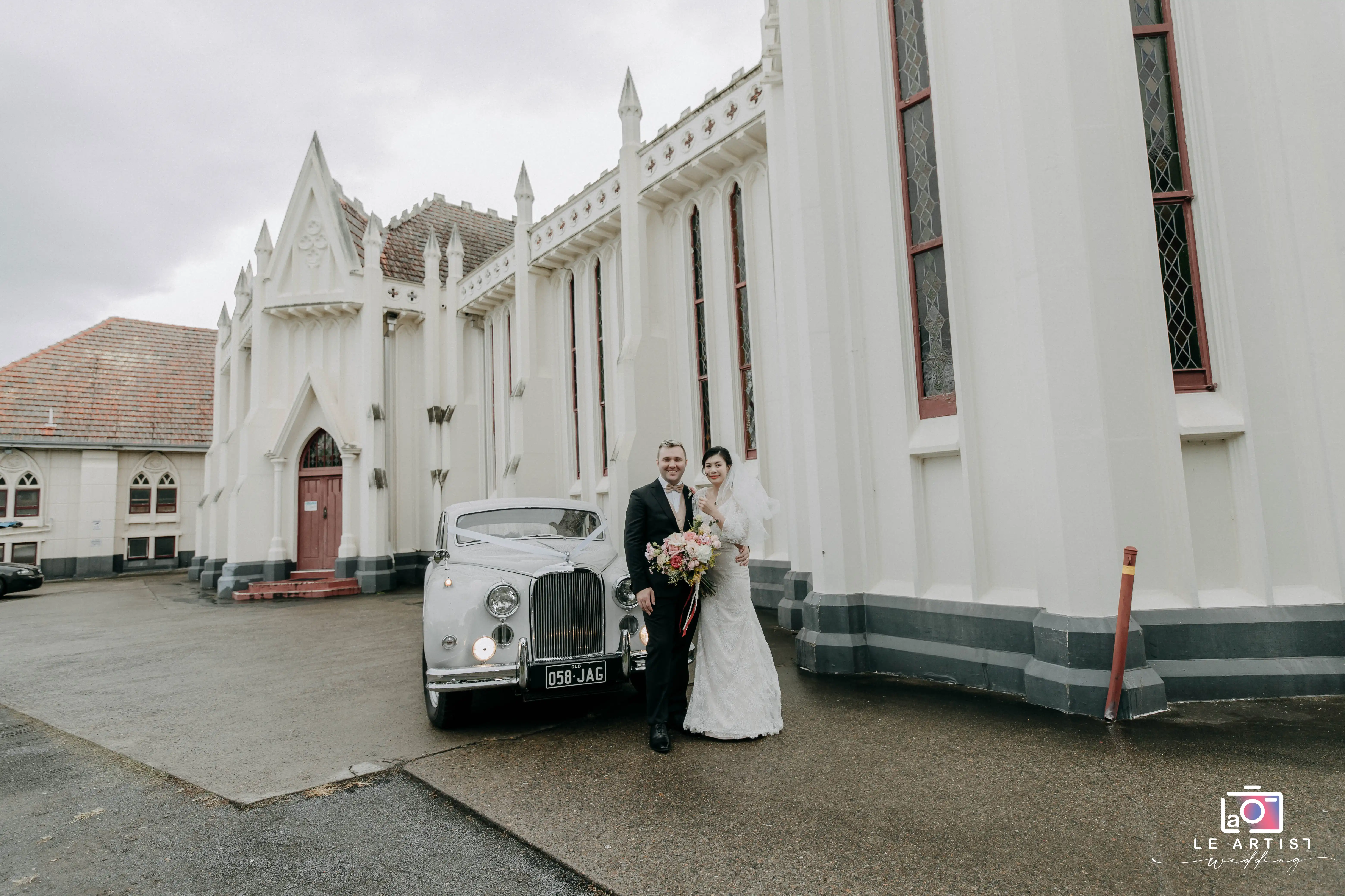 The newlyweds smile joyfully in front of a classic vintage car, with an elegant architectural backdrop enhancing the moment. Le Artist Wedding