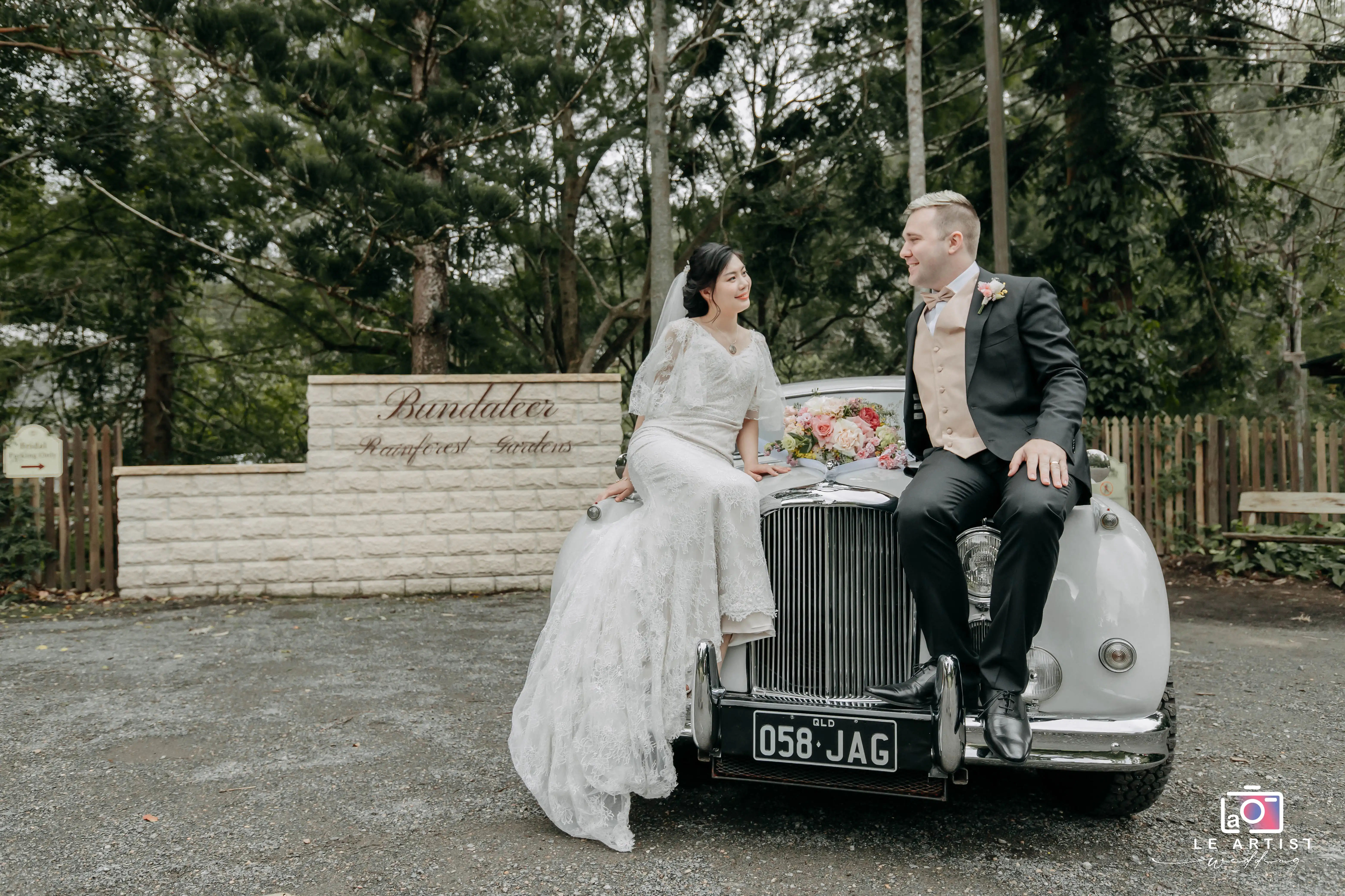 The newlyweds share a joyful moment seated on the hood of a vintage car adorned with flowers, with the beautiful backdrop of the Bandalier Rainforest Gardens. Le Artist Wedding