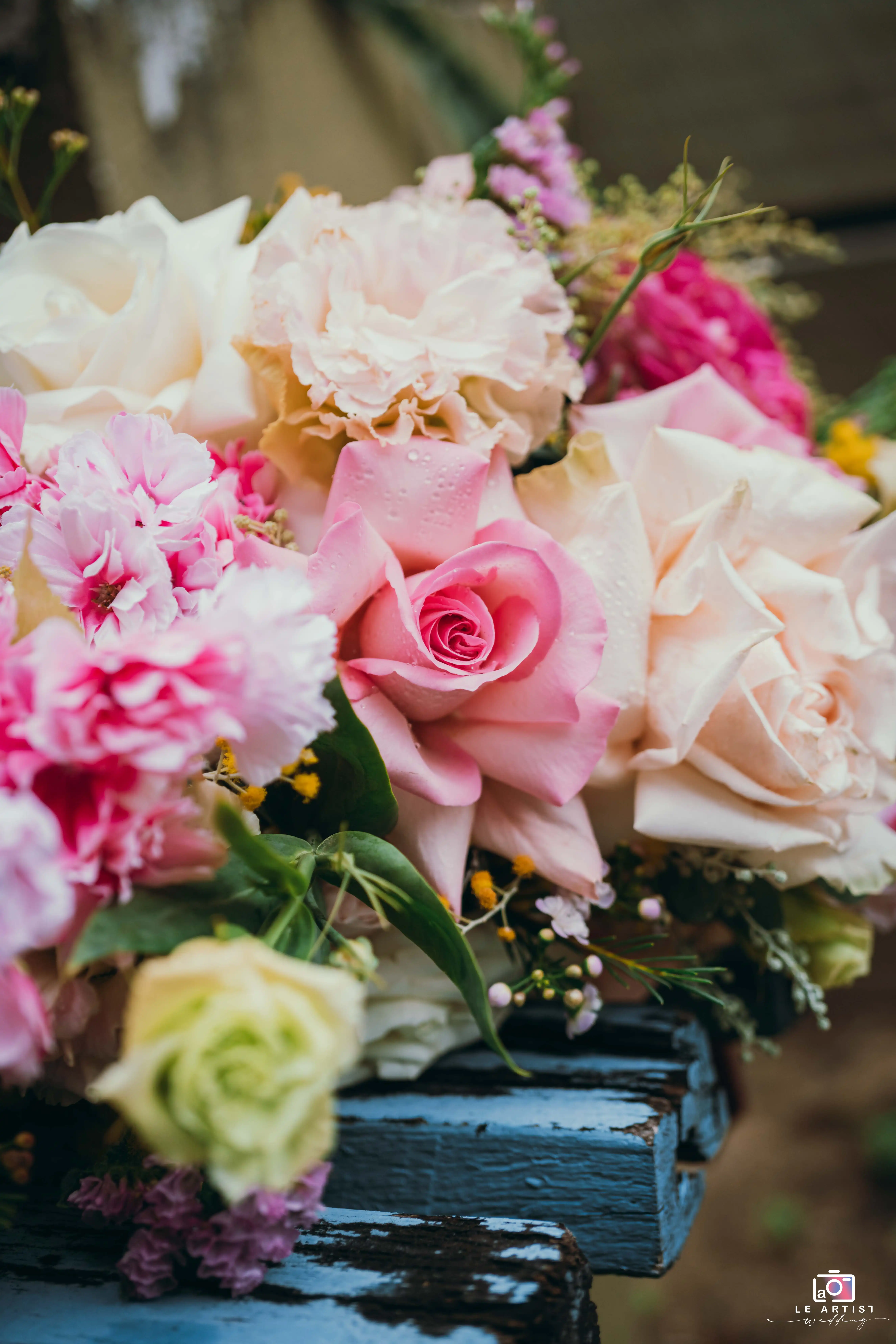 A close-up capture of a vibrant bridal bouquet showcasing a mix of pink and cream roses, accented with delicate greenery and textured flowers, resting on a rustic wooden surface. Le Artist Wedding