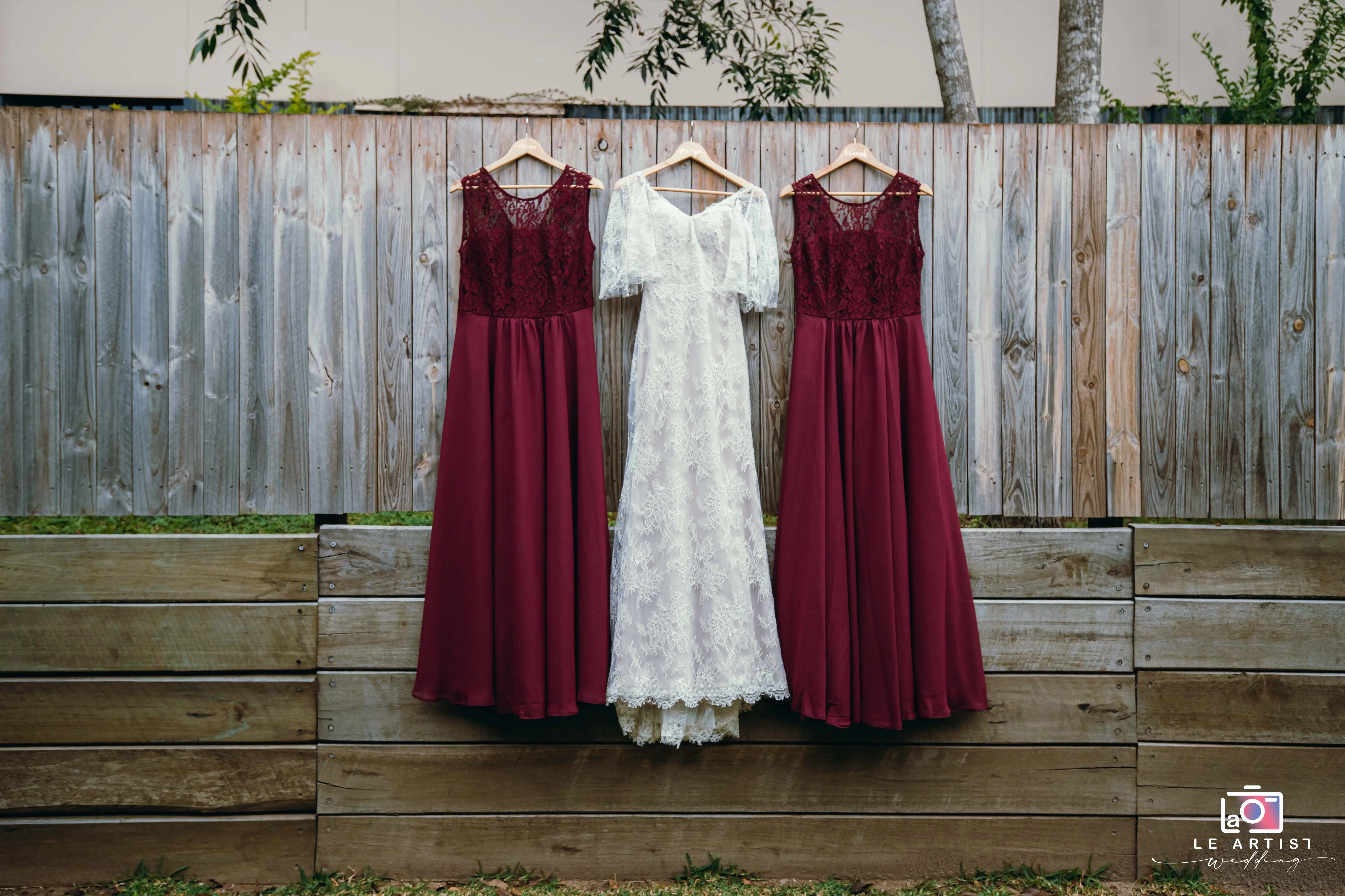 A beautifully arranged display of a bridal gown flanked by two burgundy bridesmaid dresses against a rustic wooden fence. Le Artist Wedding