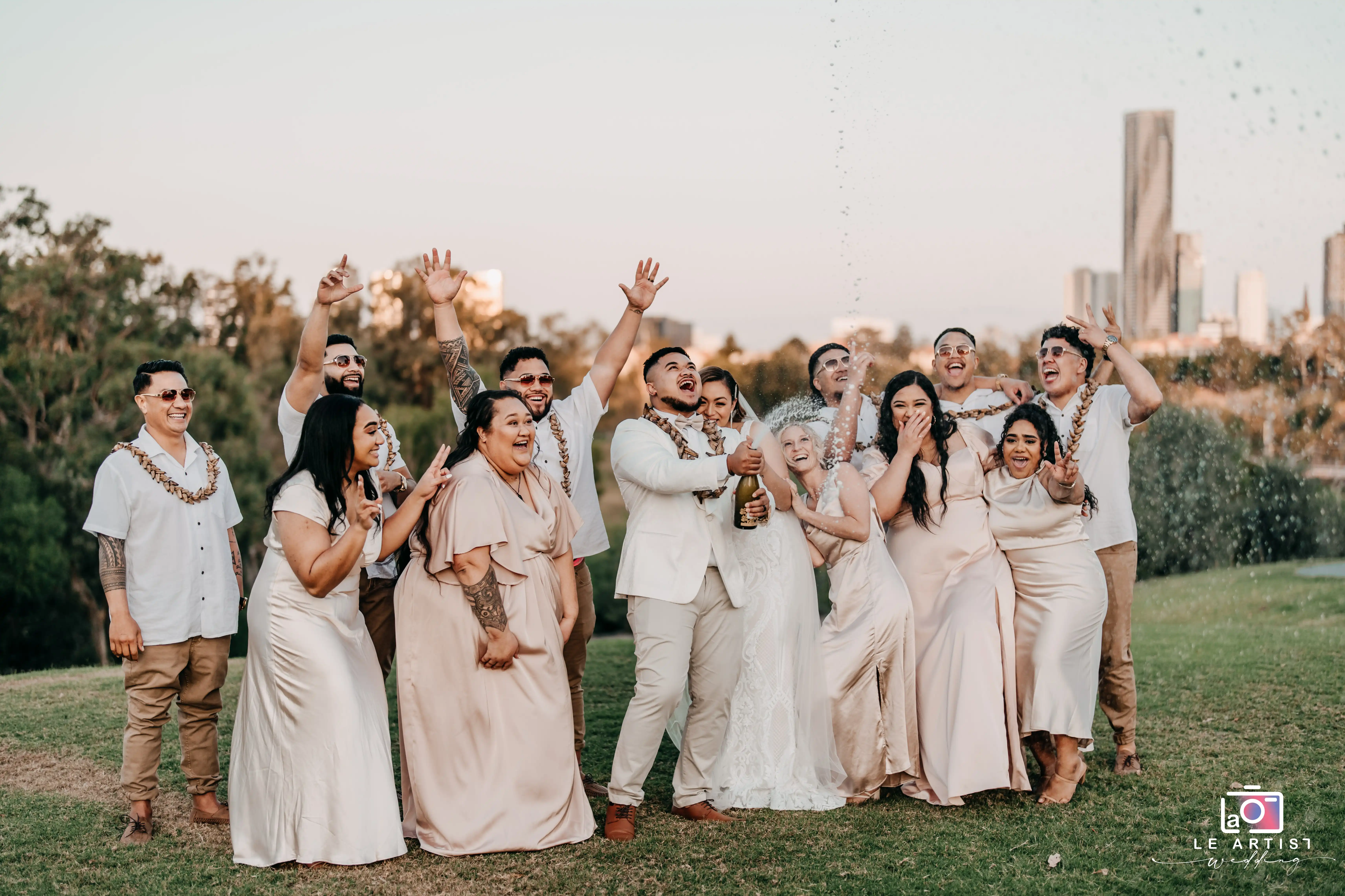 The bridal party celebrates with joy as the newlyweds pop a bottle of champagne, capturing a lively moment on the grassy hill with the skyline in the background. Le Artist Wedding