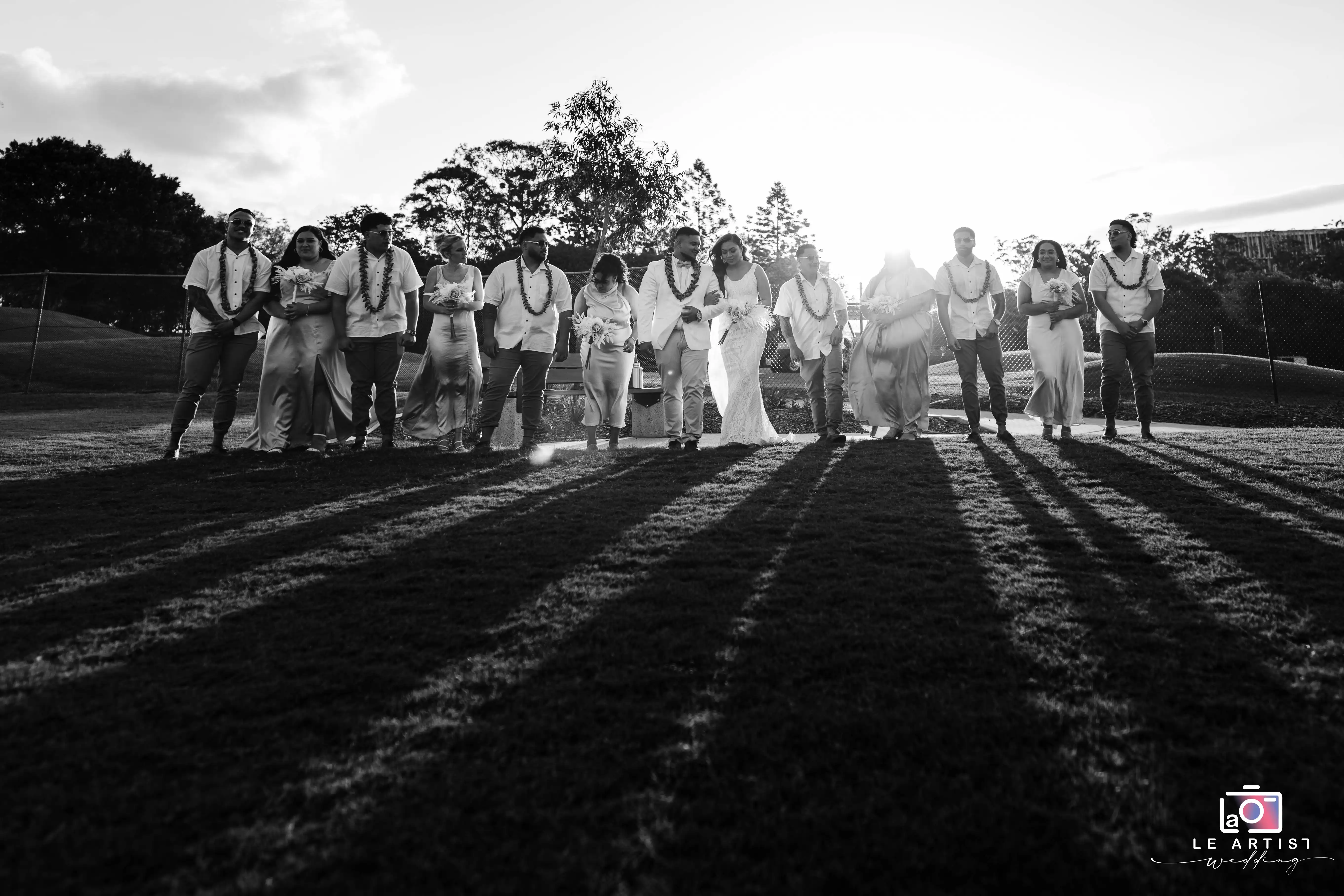 The bridal party poses together in a picturesque outdoor setting, casting long shadows as the sun sets behind them. Le Artist Wedding
