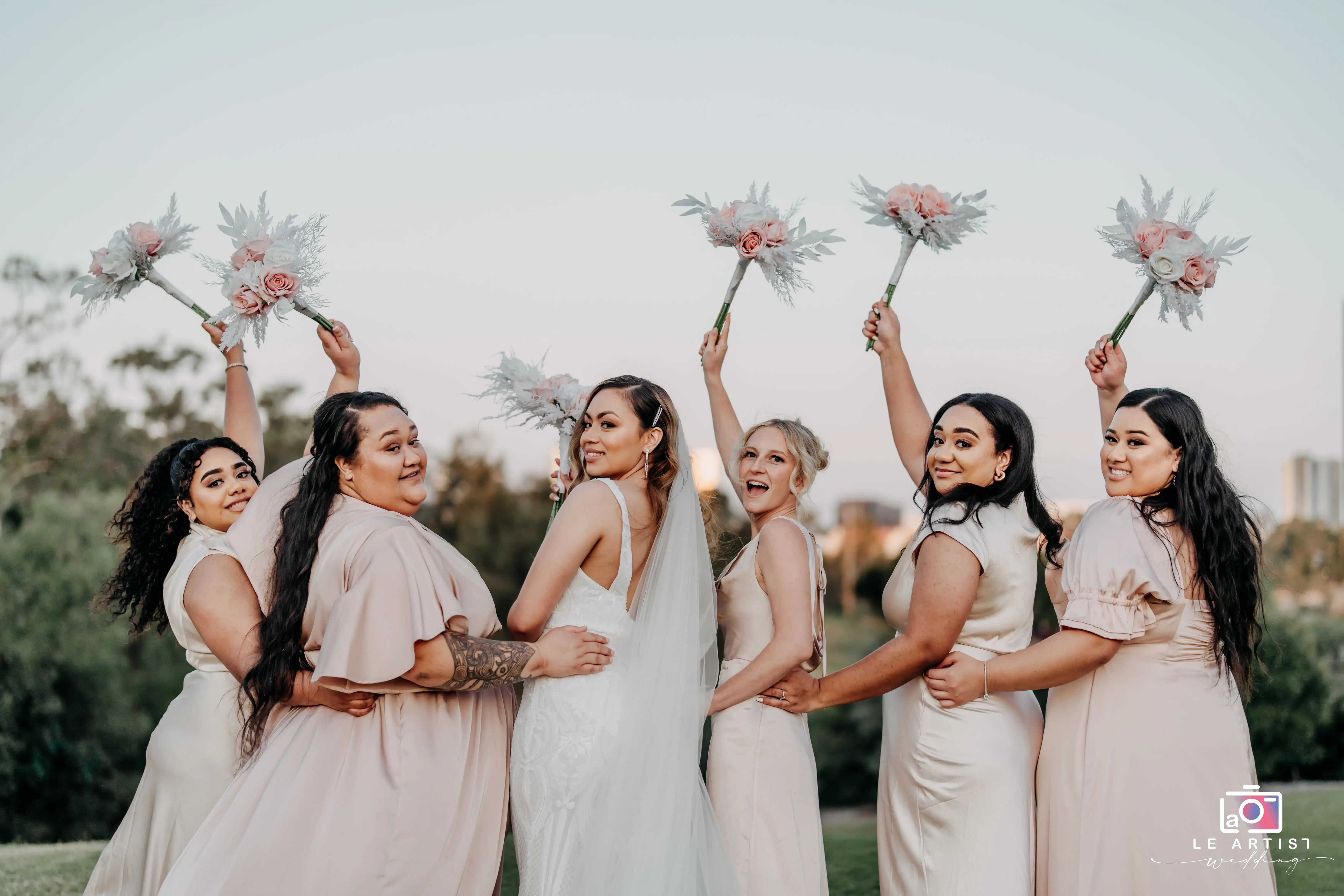 The bride and her bridesmaids strike a joyful pose, holding bouquets against a serene outdoor backdrop. Le Artist Wedding